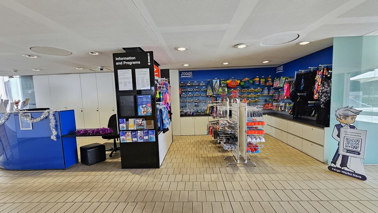 In the center is the retail section showcasing goggles, swimwear, and swimming accessories. On the left, a blue desk adorned with Christmas garland is dedicated to the Learn to Swim program, with brochures displayed in front of the black display rack. The floor is tiled with small bricks, and the ceiling features a white porous design with circular LED lights.