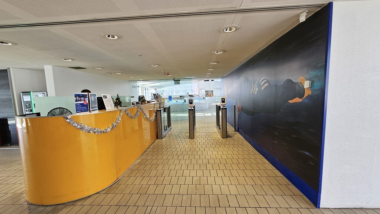 In the center stands the reception desk, featuring a caramel-brown surface embellished with Christmas garlands. To its right, a glass turnstile is in place for controlled entry. On the left, there is a spacious area with foam seating. The right wall showcases a life-sized painting of Ian Thorpe swimming underwater. The floor is tiled with small bricks, and the ceiling exhibits a white porous design with circular LED lights.