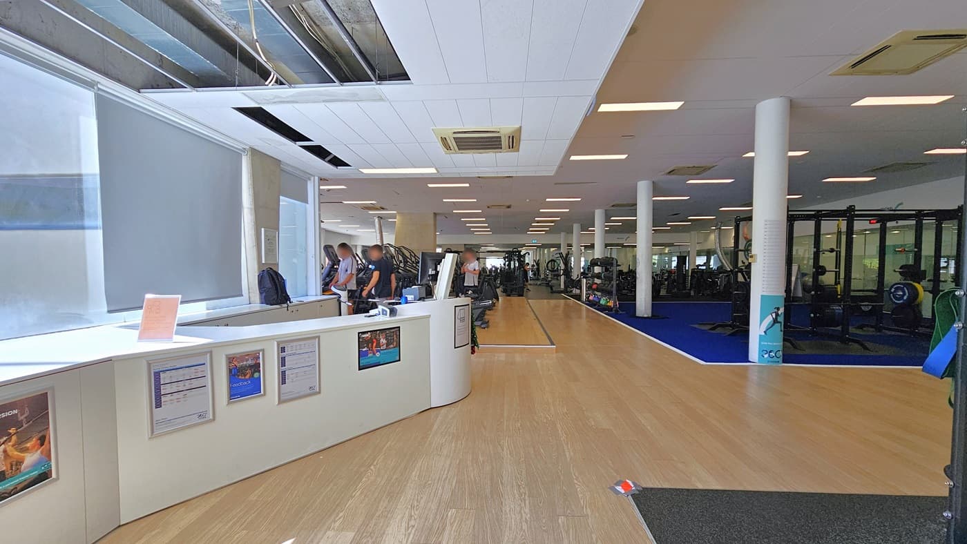 Access to the gym is through an inward-opening manual glass door. Inside, turnstiles equipped with swiping machines for access are positioned at the entry. The floor inside features a combination of black and white tiles, and the ceiling is white with centralized air conditioning. Outside, to the left, there is a lift.