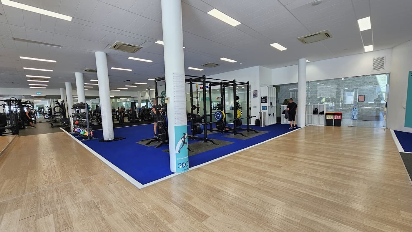 A royal blue rubber mat in the middle accommodates Smith Machines. Further in, stability balls, free weights, and additional machines are on display. To the right, there are white storage racks for belongings, and adjacent to them are two rubbish bins. The floor transitions from timber to blue rubber mats. The ceiling is white with a porous design, featuring LED lights and centralized air conditioning.