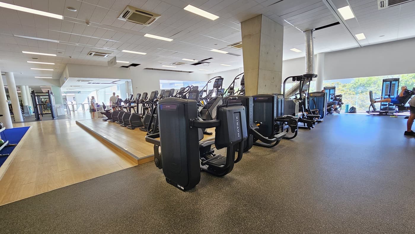 In the middle, a slightly elevated platform accommodates a lineup of cardio machines. To the left, a royal blue mat hosts stability and free weights, while further along, Smith Machines are positioned. To the right, more machines are arranged. The floor transitions from a blue rubber mat to timber in the middle and a black rubber mat on the right. The ceiling is white with a porous design, equipped with LED lights and centralized air conditioning.