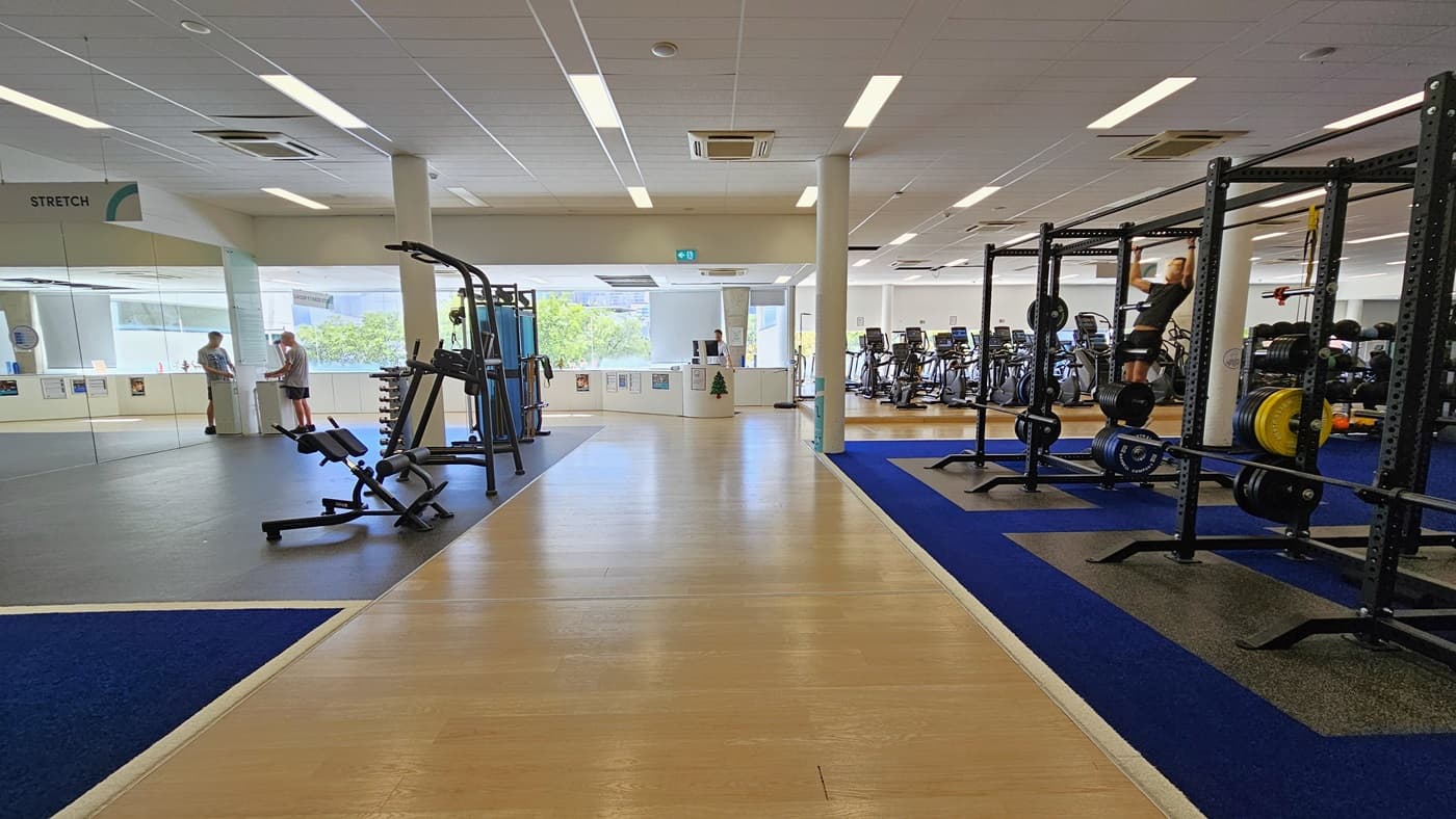 On the left is a glass door entrance with stretch machines positioned. The reception desk is in the middle. To the right, Smith machines and free weights are lined up, directly adjacent to cardio machines. The floor transitions from a blue rubber mat to a timber and a black rubber mat on the right. The ceiling is white with a porous design, featuring LED lights and centralized air conditioning. Few gym users are visible.