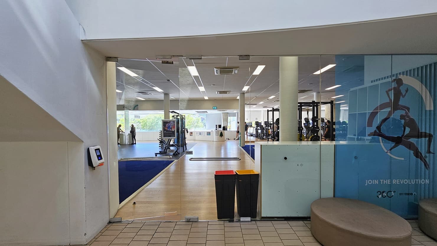 The manual inward-opening glass door serves as an alternate entrance to the gym. A swiping machine is positioned on the left for members to swipe their access cards. To the right, round cushion seatings are available. A large sports-related poster is affixed to the left glass wall. Inside, the gym's timber floor and blue mat are visible, with machines strategically positioned. Two rubbish bins are placed at the entrance. Gym users are also visible.