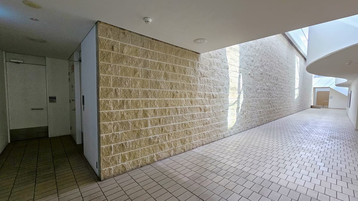 To the right is a corridor leading to the accessible bathrooms. The walls are constructed with brown bricks, and the floor is covered in brown tiles. On the left, there are two accessible bathrooms, each marked with signage indicating gender access. The ceiling is entirely white, featuring circular LED lights.