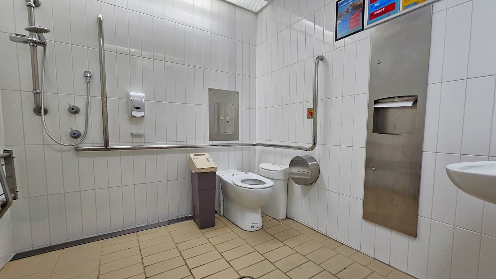 Inside the accessible bathroom. The toilet bowl, equipped with a handrail, is positioned in the right corner. Rubbish bins and a tissue dispenser are placed beside it. To the right, there is another tissue dispenser and a washbasin mounted on the wall. On the left, a shower with handrails is present, and a foldable bench is mounted on the left wall. The walls are adorned with white tiles, featuring posters affixed on top. The floor is covered in cream-coloured tiles.
