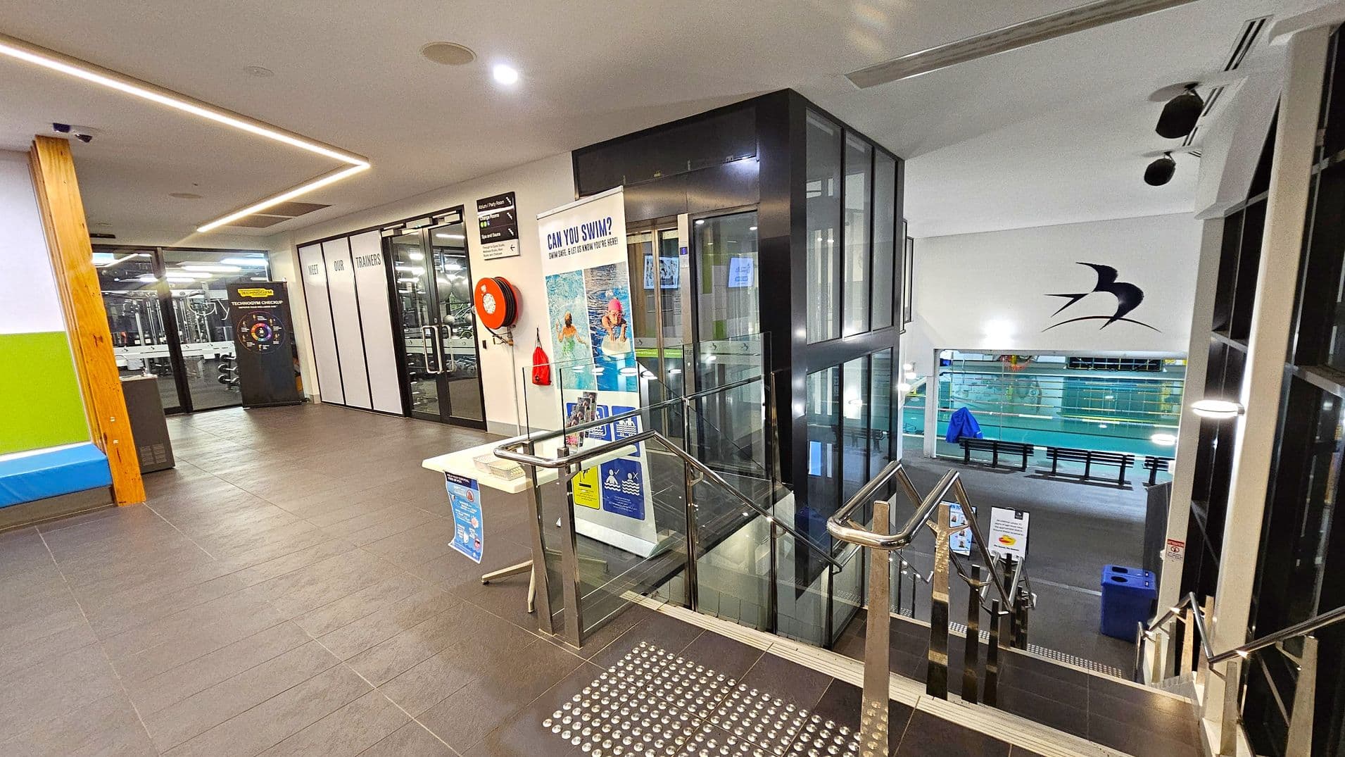 A modern indoor facility with a tiled floor. On the left, there's a gym area visible through glass doors, with a sign about trainers. A red fire hose reel is mounted on the wall. In the centre, a lift with glass panels is adjacent to a staircase with metal railings leading down. A table with informational posters is nearby. On the right, a swimming pool area is visible below, with a large bird logo on the wall. The ceiling has recessed lighting and a sleek design.