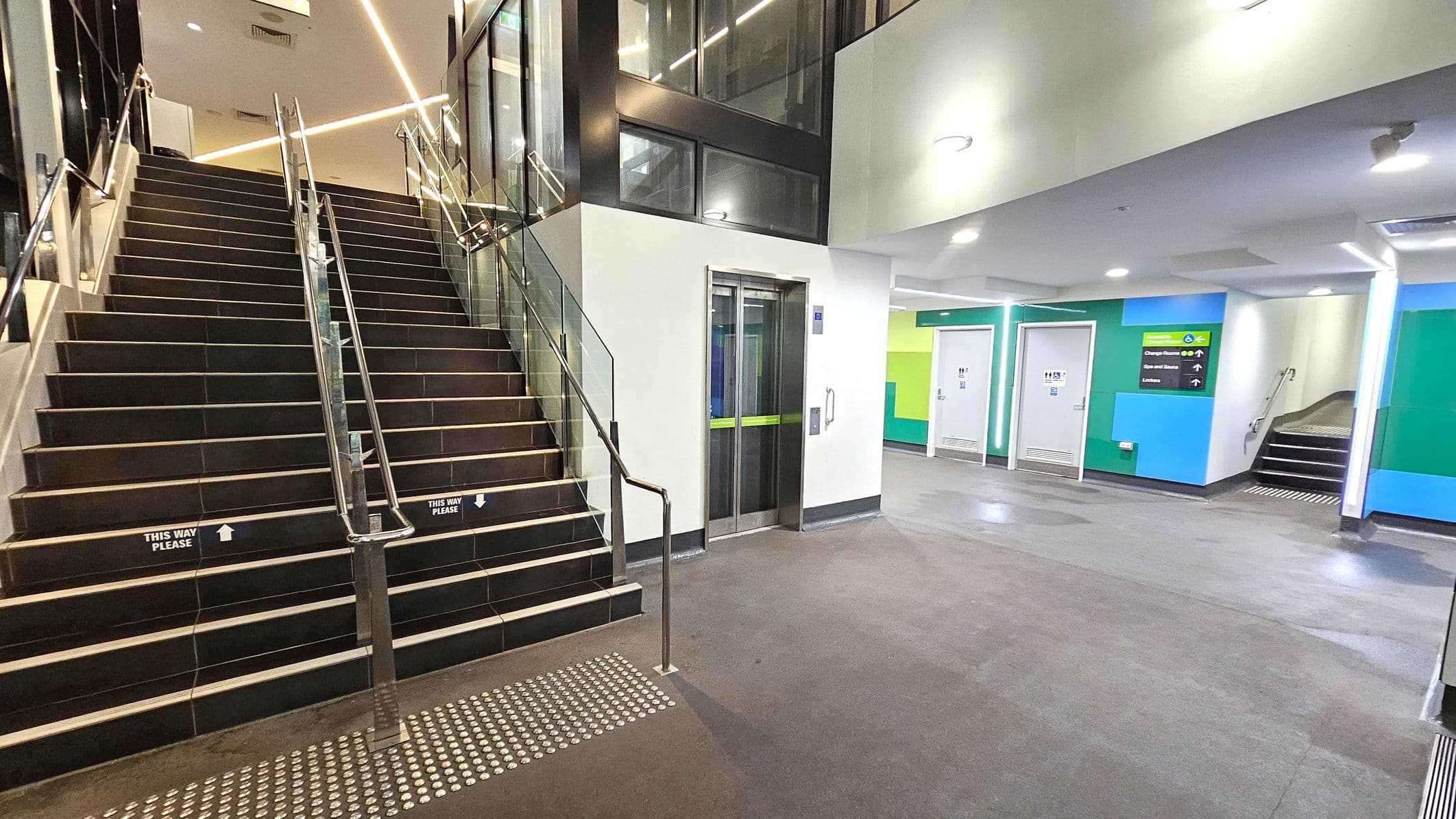 A modern indoor space with a staircase on the left featuring metal railings and glass panels. The stairs have signs reading "This Way Please." To the right, there is a lift with a green stripe on the doors. The walls are painted in shades of blue and green, with doors leading to other rooms. A sign indicates directions to changing rooms and other facilities. The floor is grey, and the ceiling has recessed lighting.