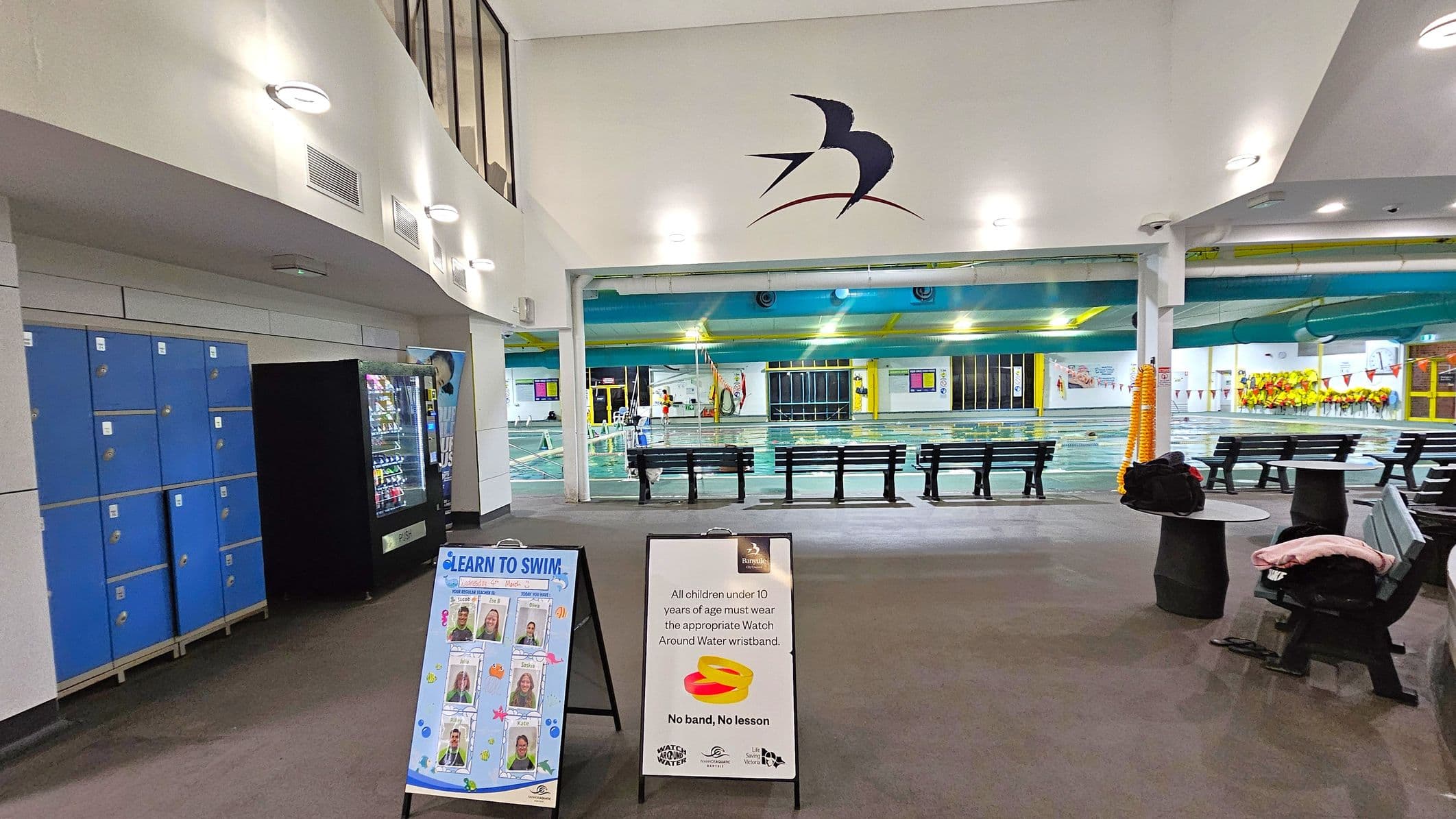 Indoor swimming pool area with blue lockers and a vending machine on the left. Two signs are in the foreground, one promoting swimming lessons and the other about wristband requirements for children. In the centre, benches face the pool, which is visible in the background. The ceiling is white with circular lights, and a bird logo is on the wall above the pool entrance. The floor is carpeted, and there are tables with clothes on the right.