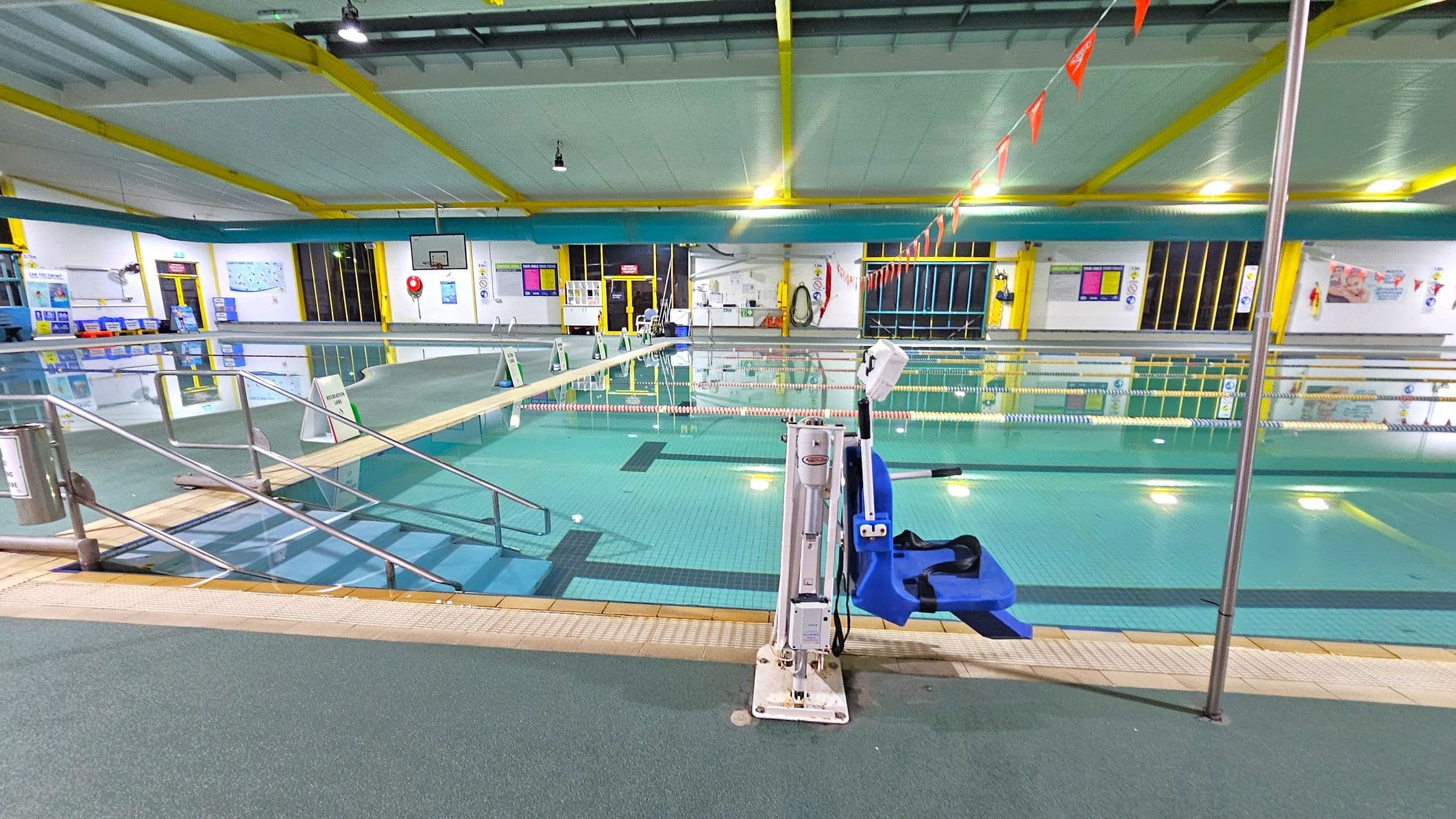 An indoor swimming pool with clear water and lane dividers. In the foreground, a blue pool lift is attached to the poolside. A ramp with metal railings leads into the water on the left. The ceiling has yellow beams and is well-lit with overhead lights. The walls are adorned with various signs and posters. A basketball hoop is visible in the background, and there are emergency equipment and doors along the walls. The floor around the pool is covered with non-slip material.