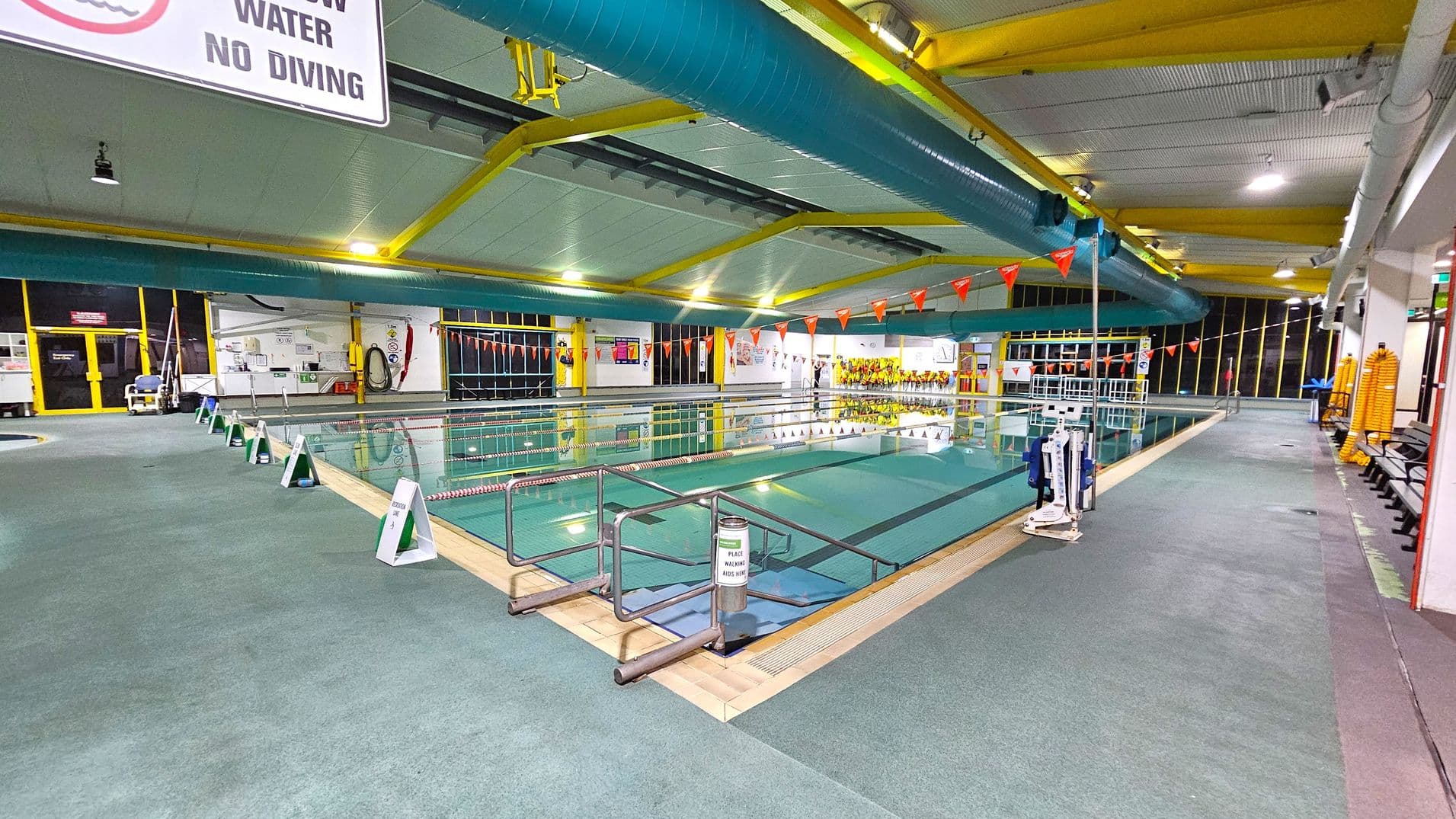 An indoor swimming pool with lanes marked by ropes and orange triangular flags overhead. The pool is surrounded by a green non-slip floor. On the left, there are signs warning of shallow water and no diving. The ceiling features yellow beams and large blue ventilation ducts. On the right, there are benches and stacked yellow flotation devices. The far end of the pool area has large windows and various safety equipment mounted on the walls.