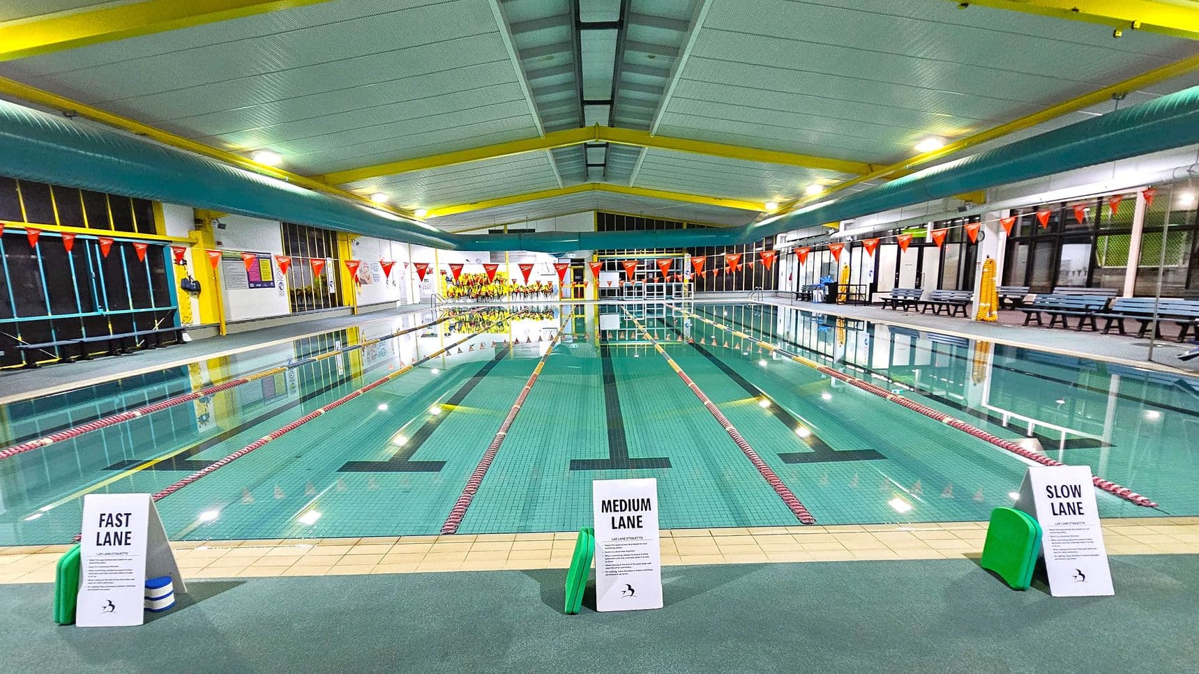 An indoor swimming pool with three lanes marked by signs: "Fast Lane" on the left, "Medium Lane" in the centre, and "Slow Lane" on the right. The pool is bordered by red and white lane dividers. The ceiling is white with yellow beams, and the walls have large windows and benches along the sides. Orange triangular flags hang above the pool. The floor around the pool is tiled, and there are green kickboards near each lane sign.