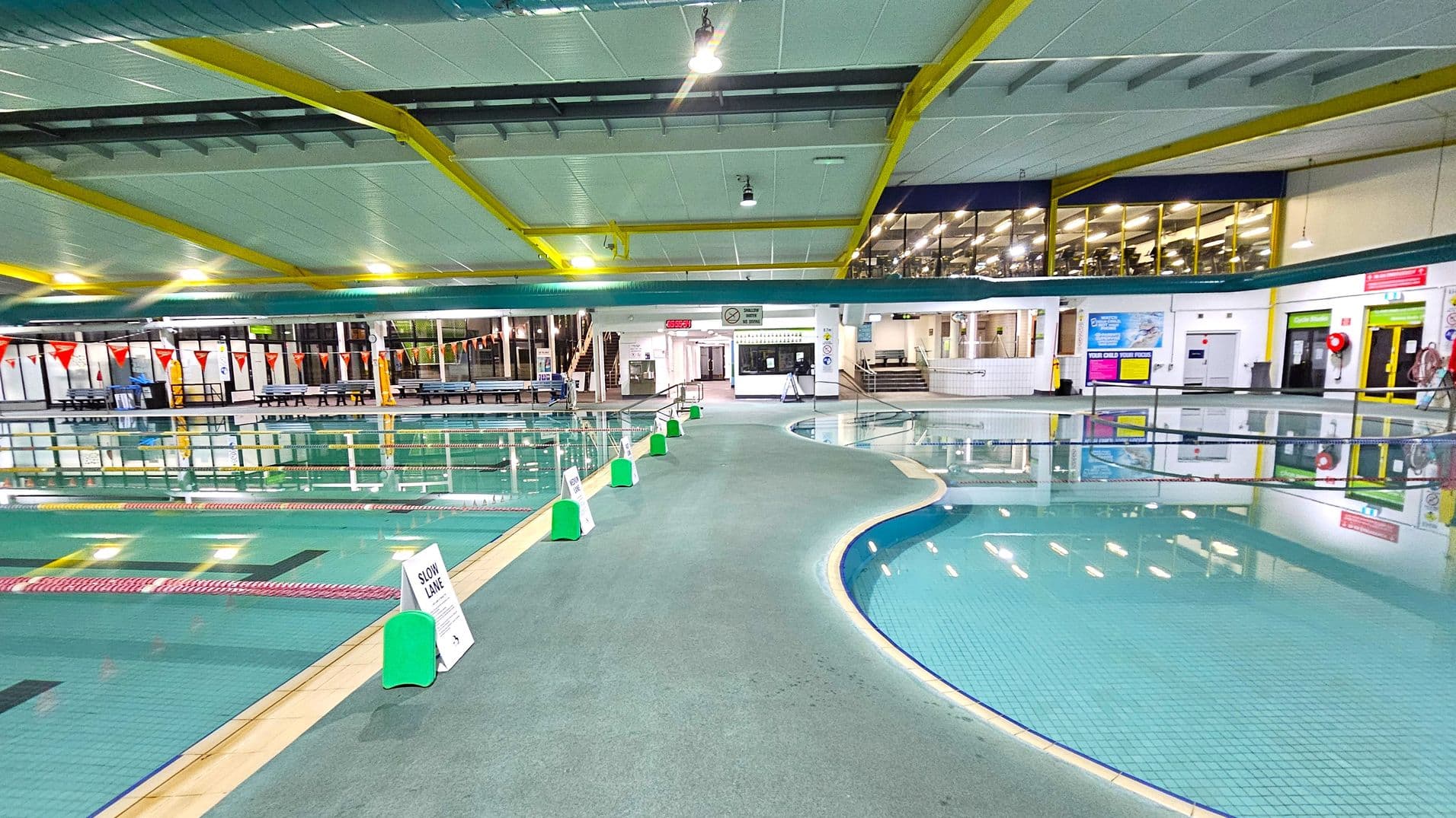 Indoor swimming pool area with a wide walkway in the centre, bordered by pools on both sides. The left side features a lap pool with lane dividers and triangular flags above. The right side has a smaller, shallow pool. Green and white signs are placed along the walkway. The background shows a glass-walled gym area and various doors and signs. The ceiling is white with yellow beams and overhead lights.
