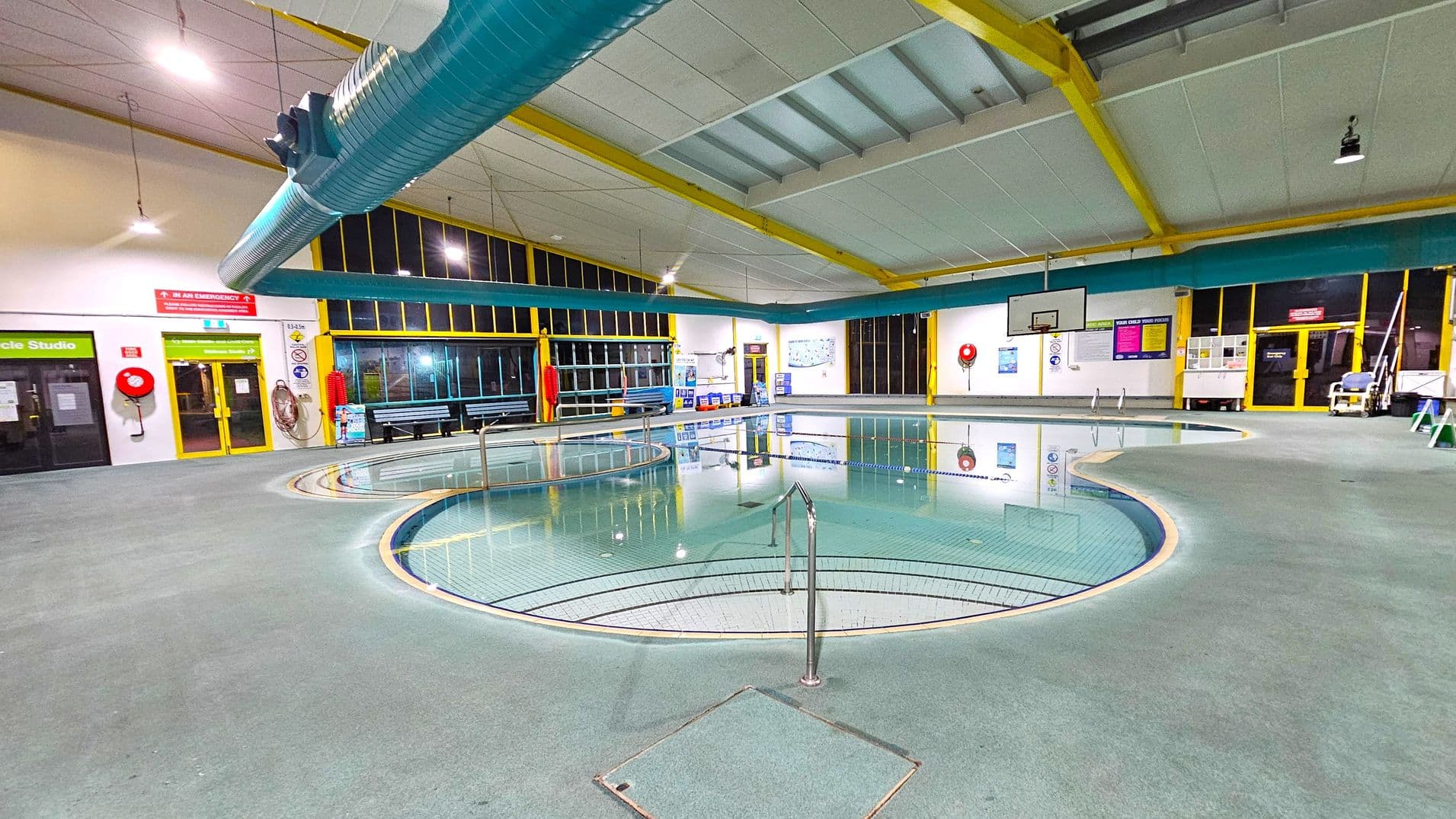 Indoor swimming pool area with a circular pool in the foreground, surrounded by a grey tiled floor. A metal handrail leads into the pool. Large windows with yellow frames line the walls, and a blue ventilation duct runs across the ceiling. On the left, there are doors labelled "Cycle Studio" and emergency equipment like a fire hose. On the right, a basketball hoop is mounted on the wall. Various signs and safety equipment are visible around the pool area.