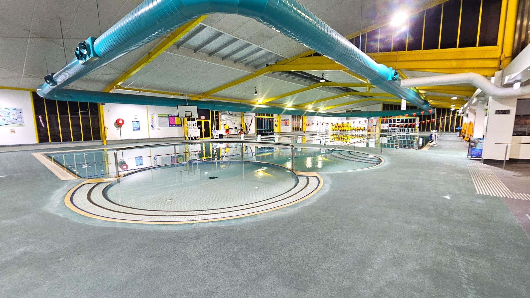 Indoor swimming pool area with a circular shallow pool in the foreground. The floor is grey and textured. In the background, a larger pool is visible with lane dividers and a basketball hoop. The ceiling is supported by yellow beams, and large blue ventilation ducts run across it. Walls have windows with yellow frames and various posters. On the right, there are shelves with equipment and a walkway leading further into the facility.