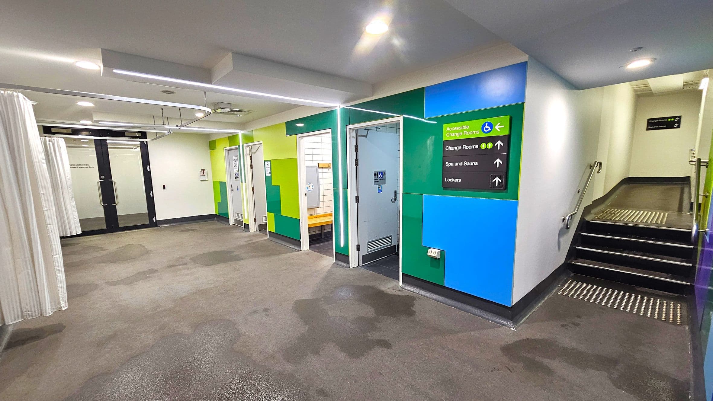 A corridor with a carpeted floor and a white ceiling featuring recessed lighting. On the left, there are glass doors with curtains. In the centre, there are several doors leading to changing rooms, with colourful green and blue walls. A sign indicates directions to accessible change rooms, spa and sauna, and lockers. On the right, a staircase with a handrail leads upwards.