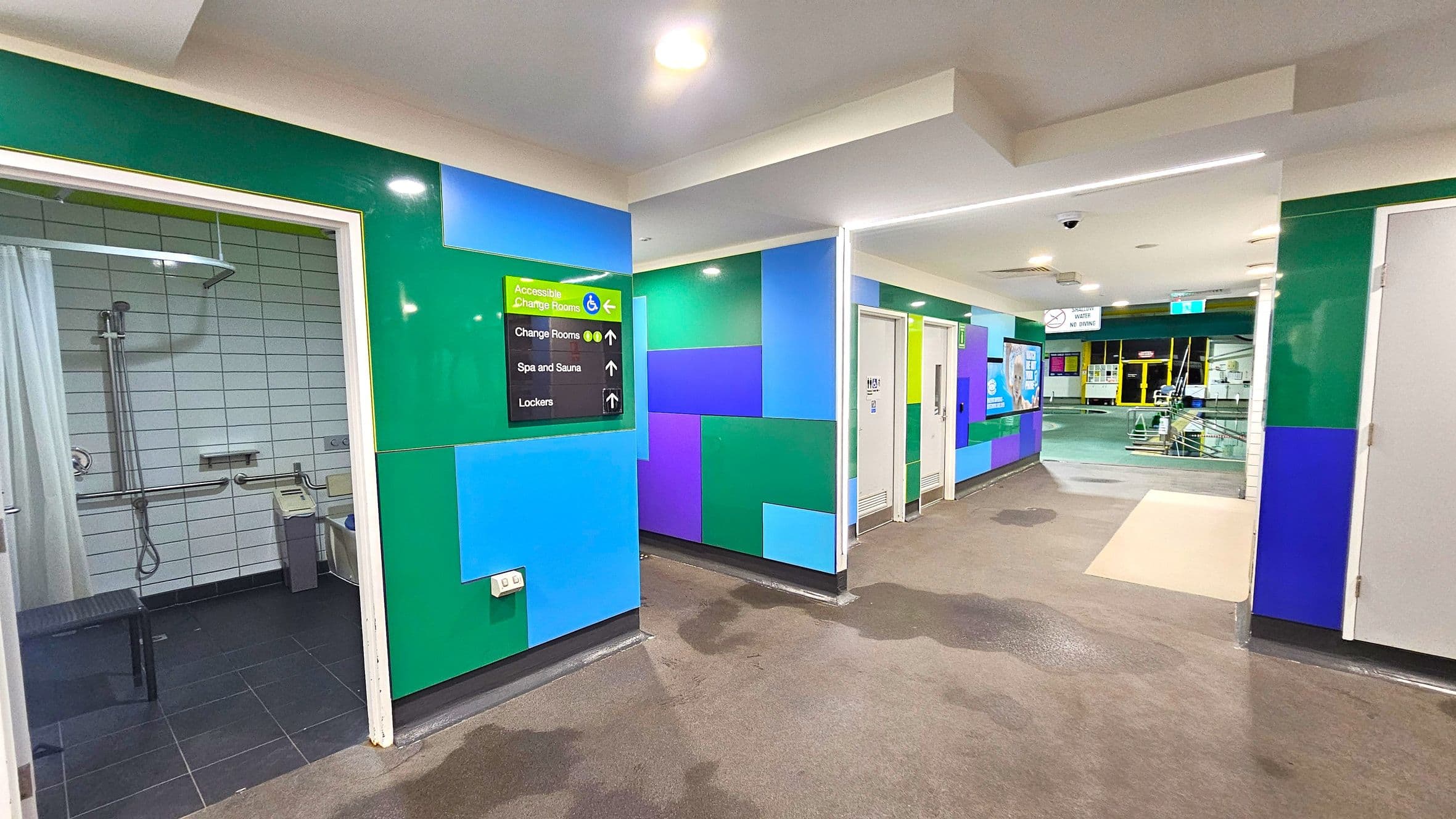 On the left, there is an accessible shower room with a curtain, grab bars, and a showerhead. The walls are tiled in white. In the centre, a colourful wall with green, blue, and purple panels features a sign indicating directions to accessible change rooms, spa and sauna, and lockers. On the right, there are doors leading to other facilities. The floor is dark with some wet patches, and the ceiling is white with recessed lighting. In the background, there is a view of a pool area.