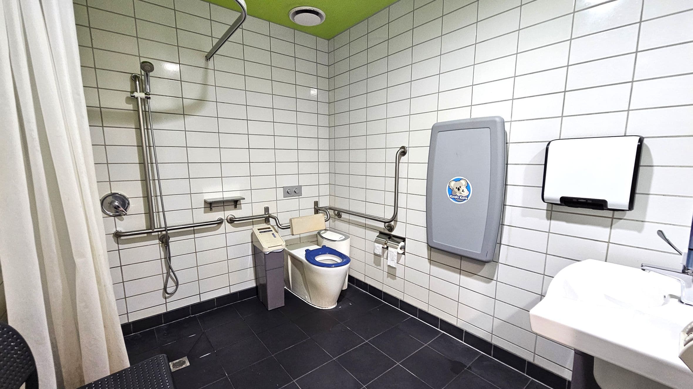 A public accessible bathroom with white tiled walls and a green ceiling. On the left, there is a shower area with a curtain and a handheld showerhead attached to a vertical rail. In the centre, a toilet with a blue seat is equipped with support rails and a small control panel. On the right, a baby changing station is mounted on the wall above a paper towel dispenser. A white sink with a lever tap is visible in the foreground. The floor is covered with dark tiles.
