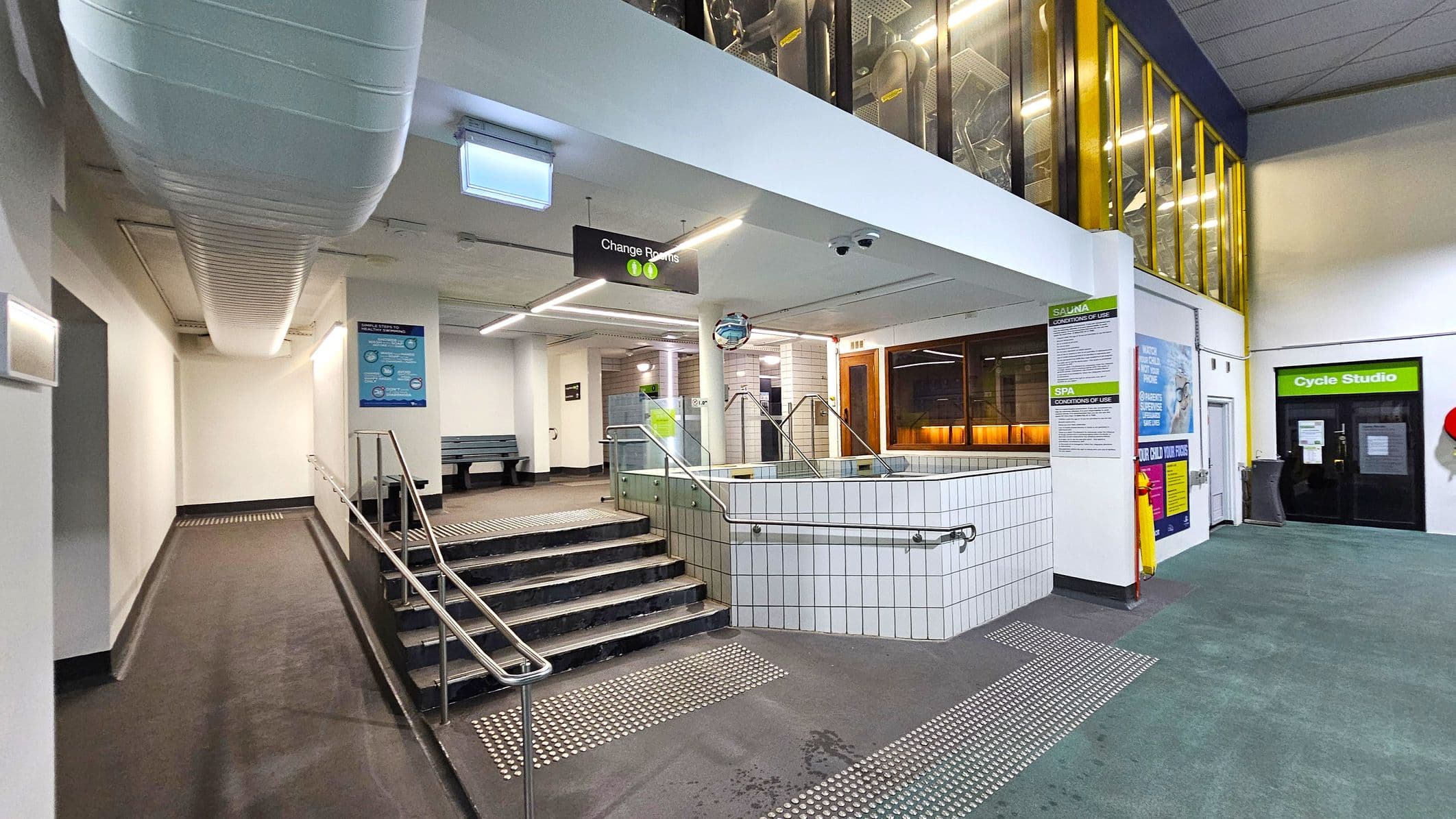 A modern indoor facility with a tiled hot tub in the centre, surrounded by metal railings. To the left, a ramp and stairs lead to a corridor with a bench. Overhead, a large white duct runs along the ceiling. On the right, a glass-walled area with yellow frames and a door labelled "Cycle Studio". Signs for changing rooms and spa conditions are visible. The floor is a mix of textured tiles and carpet, and the ceiling is fitted with fluorescent lights.