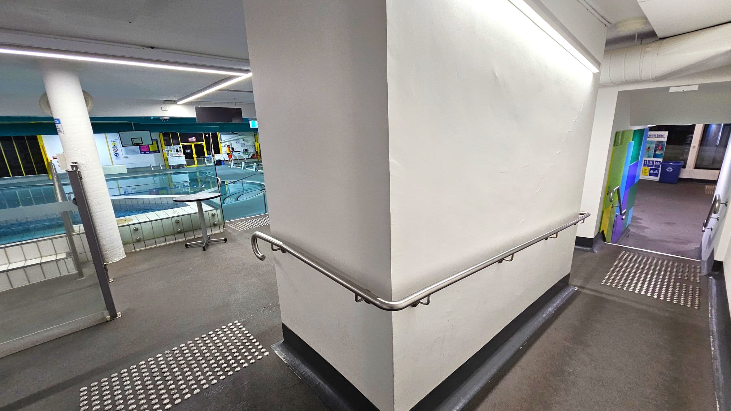 A corridor with a white wall in the centre, featuring a metal handrail. On the left, there's a view of an indoor swimming pool with glass barriers and a small round table. The pool area has yellow-framed windows and various signs. On the right, a ramp with tactile paving leads downwards, with colourful wall panels visible at the bottom. The ceiling has linear lighting, and the floor is dark with tactile indicators.