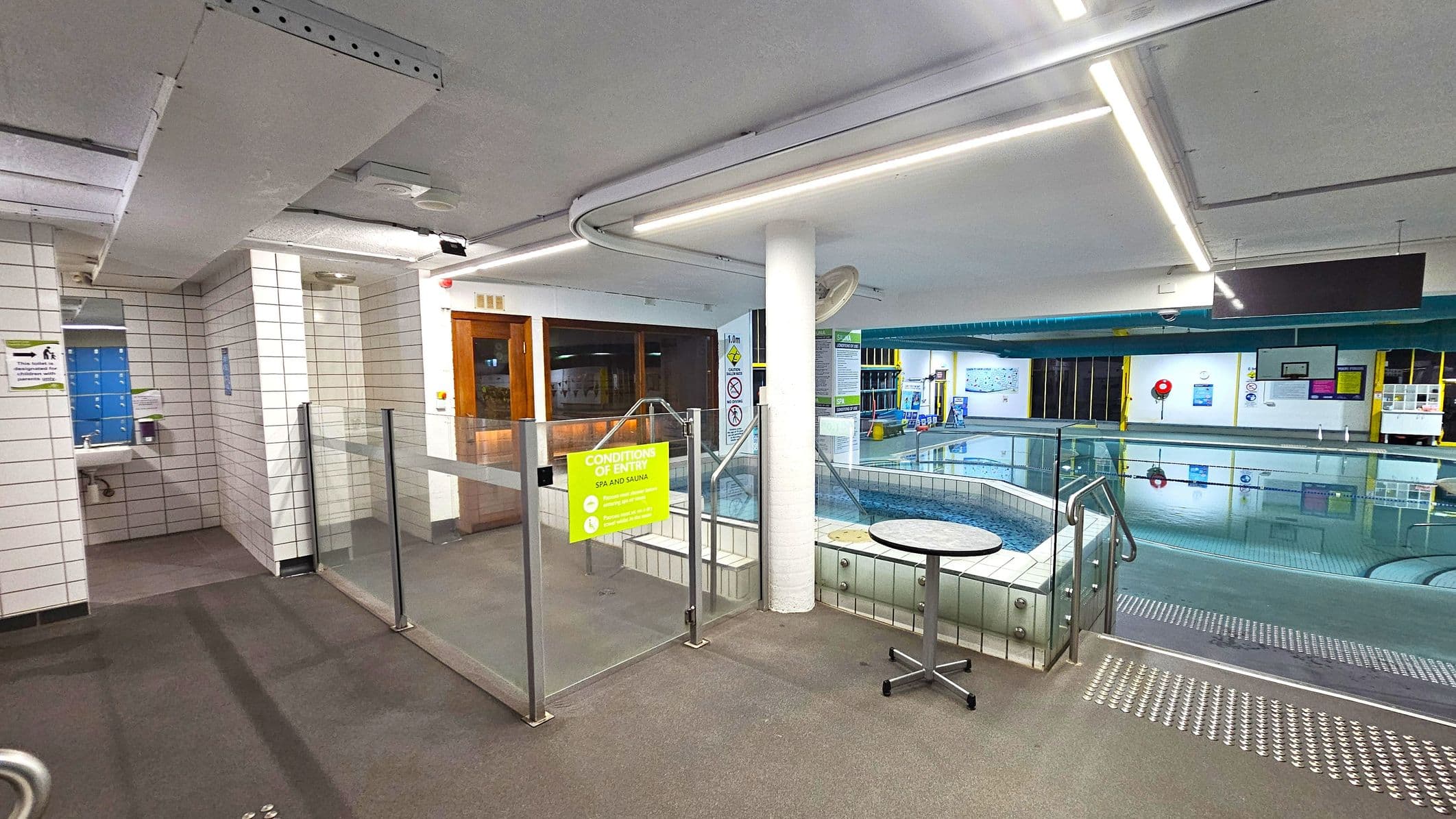 A swimming pool area with a tiled floor and ceiling. On the left, there is a sink and mirror next to a tiled wall. In the centre, a glass barrier with a yellow sign reading "Conditions of Entry" separates the pool area. A small round table is nearby. On the right, the pool is visible with clear water, surrounded by more tiled surfaces. The background shows various signs and equipment, with a lifebuoy on the wall. The ceiling has fluorescent lighting.