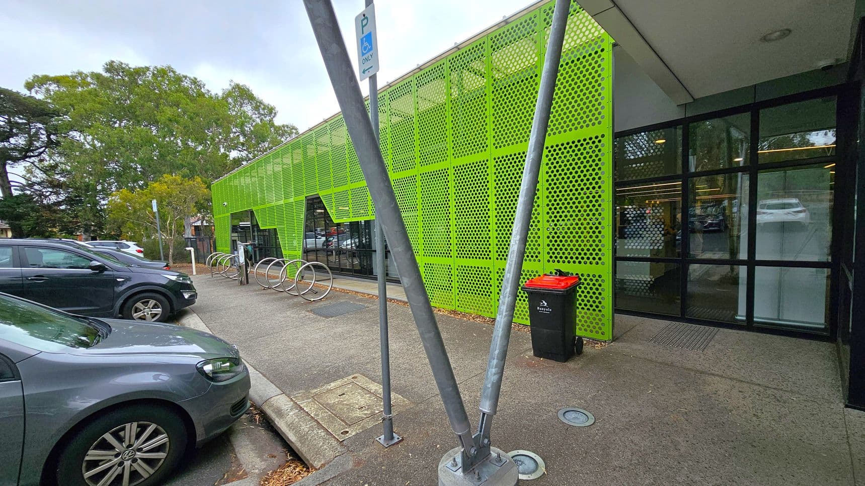 A modern building with a bright green perforated metal facade on the right. Large glass windows are visible beneath the green panels. In front, there are metal bike racks and a black bin with a red lid. Two metal poles are positioned in the foreground. On the left, several parked cars are visible, and trees are in the background. The ground is paved, and the ceiling of the building is flat and white.