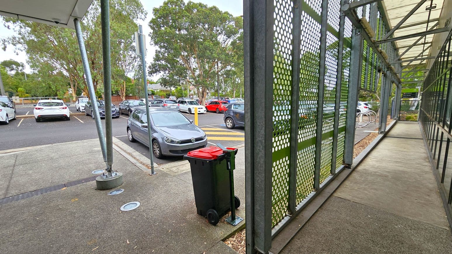 A parking area with several cars is visible on the left, surrounded by trees. A grey car is parked near a red-lidded rubbish bin on a paved surface. On the right, a walkway is enclosed by a metal structure with perforated panels and a transparent roof. The ground is a mix of concrete and green patches.