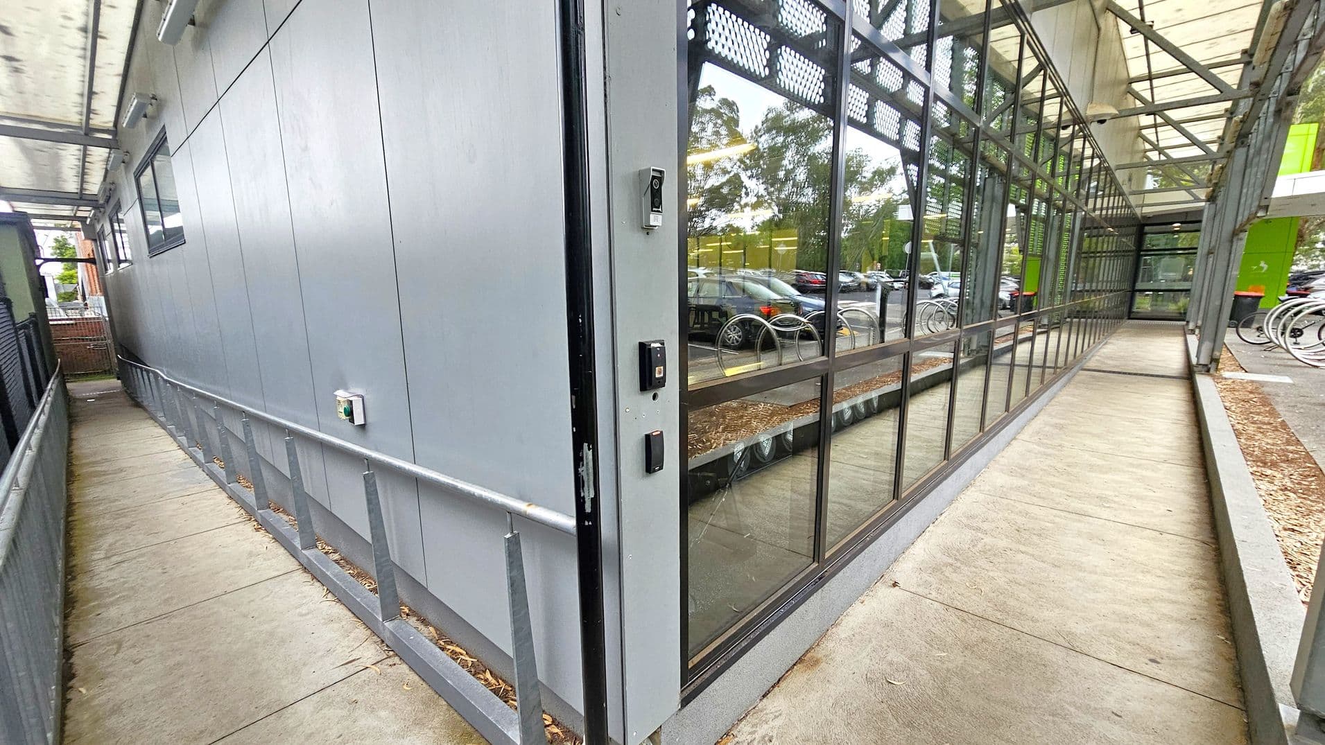 A narrow walkway with a metal railing on the left, adjacent to a grey building wall with a small window. On the right, a glass wall reflects parked cars and trees. The path is covered by a transparent roof supported by metal beams. The floor is concrete, and there are security devices mounted on the wall near the glass section. In the background, bicycles are parked near a green structure.