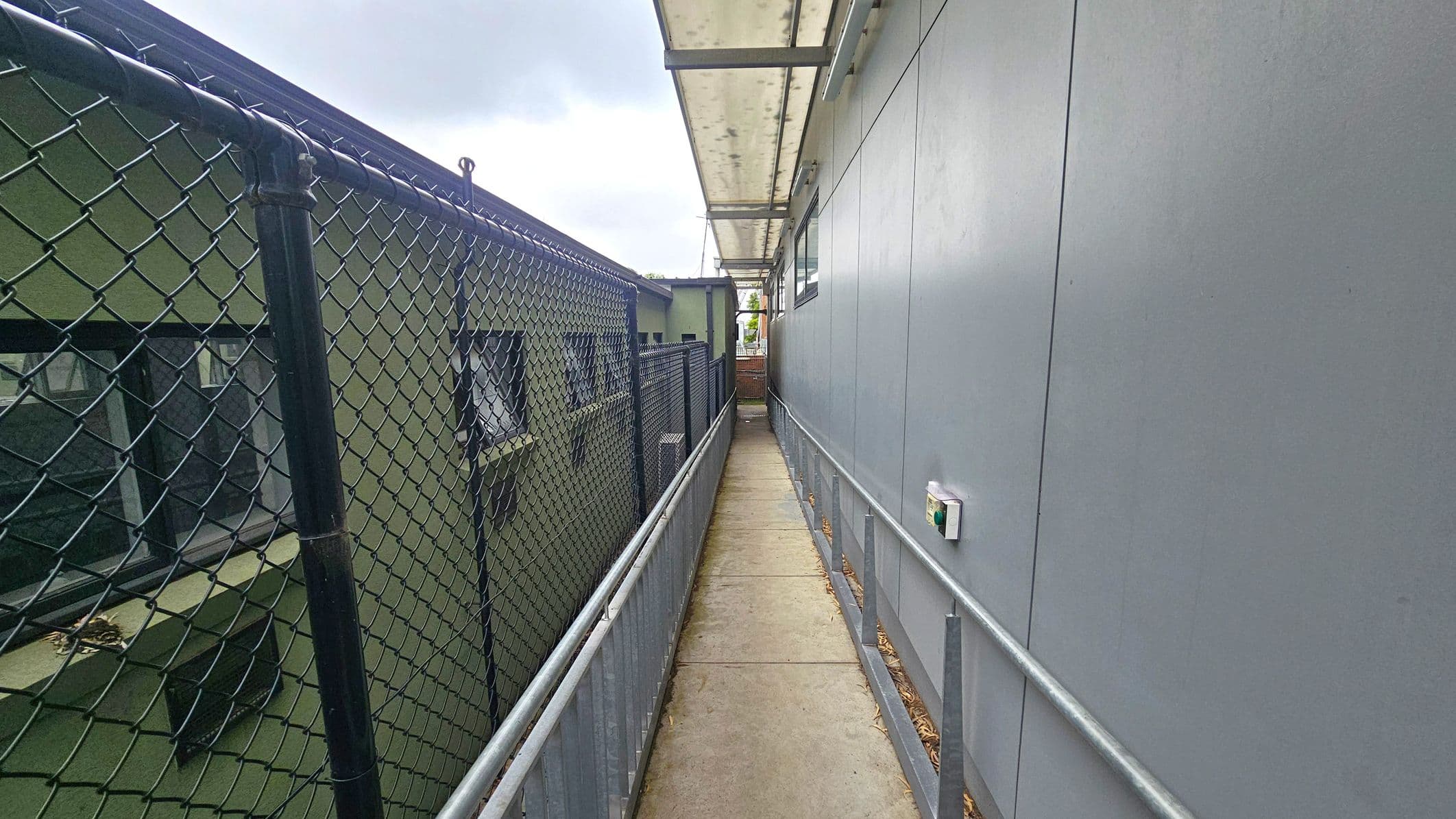 A narrow walkway with a concrete floor runs between two structures. On the left, there's a green building with windows, separated by a chain-link fence. On the right, a grey building with a smooth wall and a small device attached. Above, a translucent canopy provides cover. The walkway is bordered by metal railings on both sides.