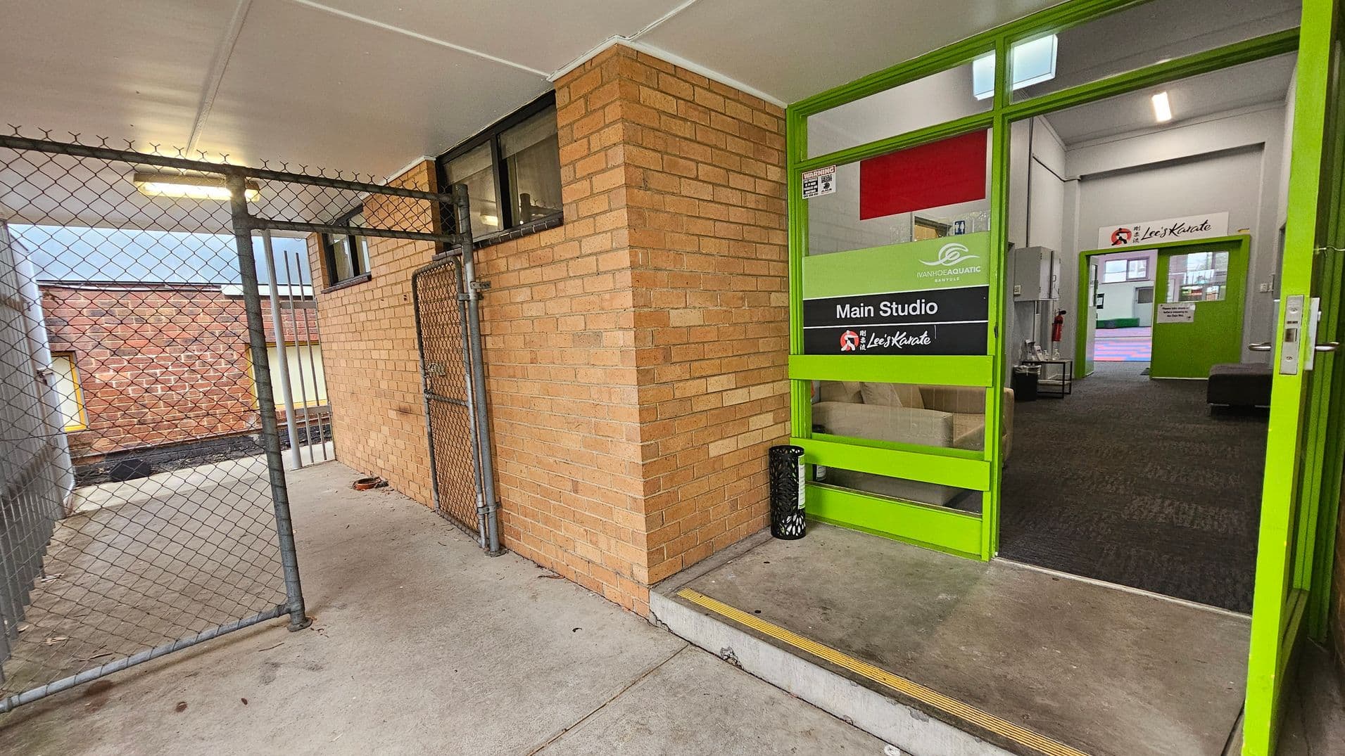 A corridor with a chain-link fence on the left and a brick wall on the right. The floor is concrete. The entrance to a building is on the right, featuring a green-framed glass door with signs for "Main Studio" and "Lee's Karate." Inside, the carpeted floor is visible, along with a fire extinguisher and a small table. The ceiling is white with fluorescent lighting.