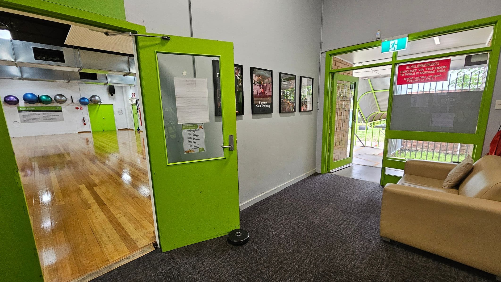 A room with bright green doors and a grey carpeted floor. On the left, an open door leads to a wooden-floored studio with exercise balls on the wall. On the right, another green-framed door opens to an outdoor area with grass visible. A beige sofa with a cushion is in the foreground. Several framed posters are mounted on the wall between the doors. An emergency exit sign is above the right door.
