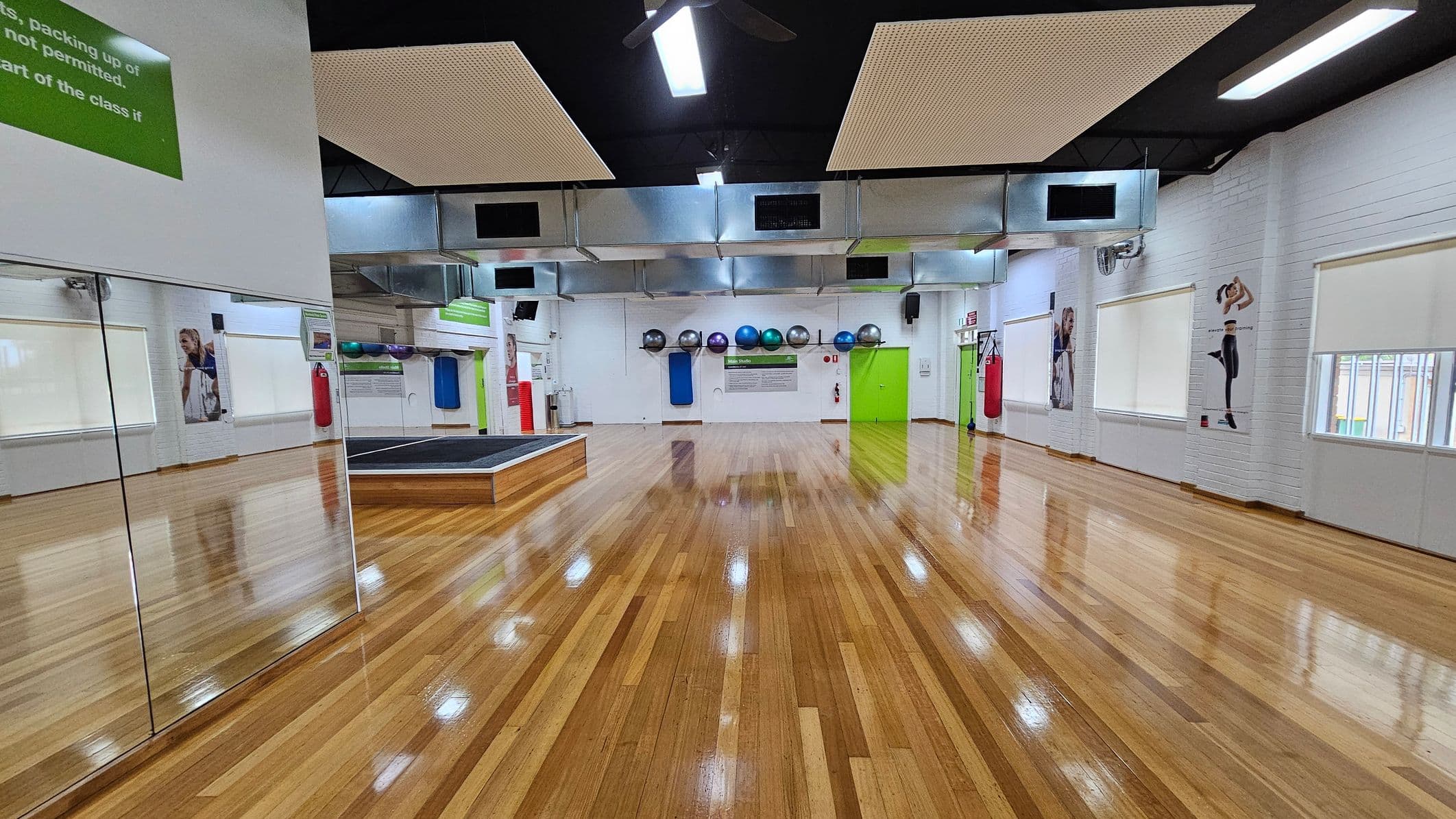A spacious gym room with polished wooden floors. On the left, there are large mirrors reflecting the room. The back wall features exercise balls in various colours on a rack, a green door, and a small stage. The right wall has posters of people exercising and several windows with blinds. The ceiling is black with rectangular light fixtures and ventilation ducts. A red punching bag hangs near the back wall.