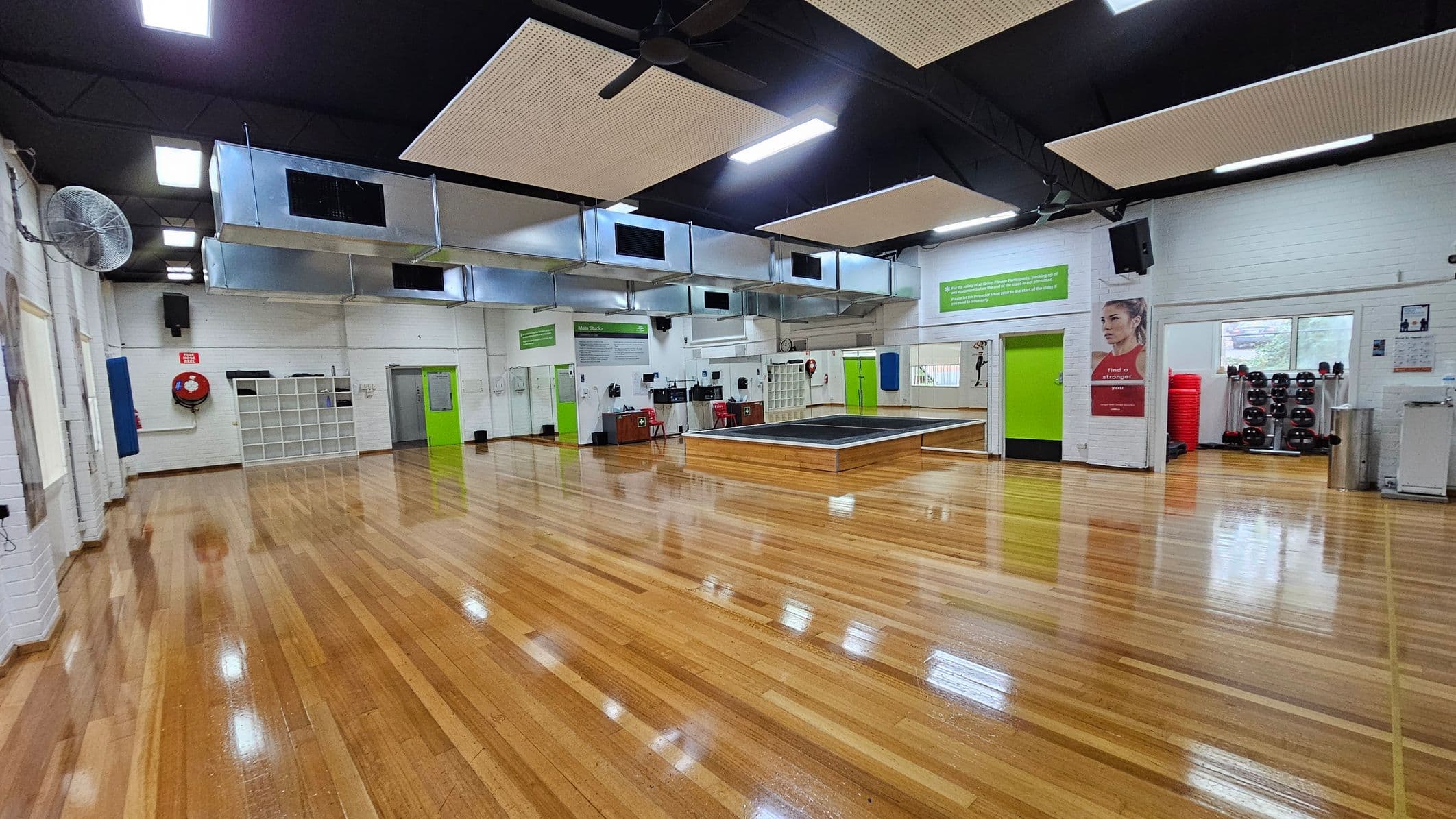 A spacious gym studio with polished wooden floors and a high ceiling featuring large ventilation ducts and ceiling fans. On the left, there are white brick walls with a fire hose reel and shelving units. In the centre, a raised platform is visible. On the right, a green door is flanked by posters and a window. Exercise equipment, including weights and stacked mats, is neatly arranged. Bright overhead lights illuminate the room.