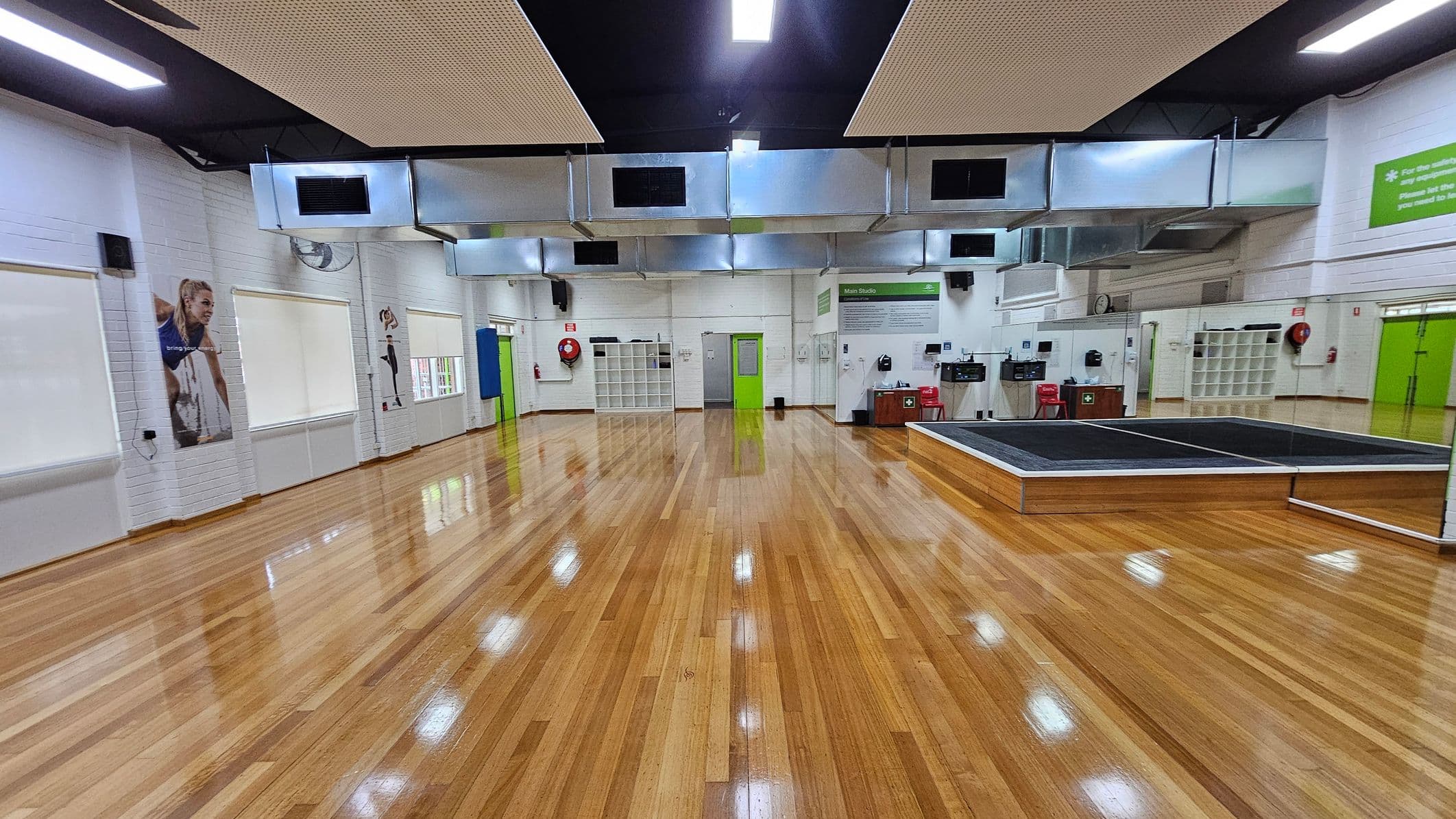 A spacious room with polished wooden floors and white brick walls. On the left, there are large windows with blinds and posters of athletes. The centre features a green door and a wall with cubbyholes. On the right, there's a raised platform with mirrors and a green sign above. The ceiling has large metallic air ducts and rectangular lights. Red chairs and fire safety equipment are visible along the walls.