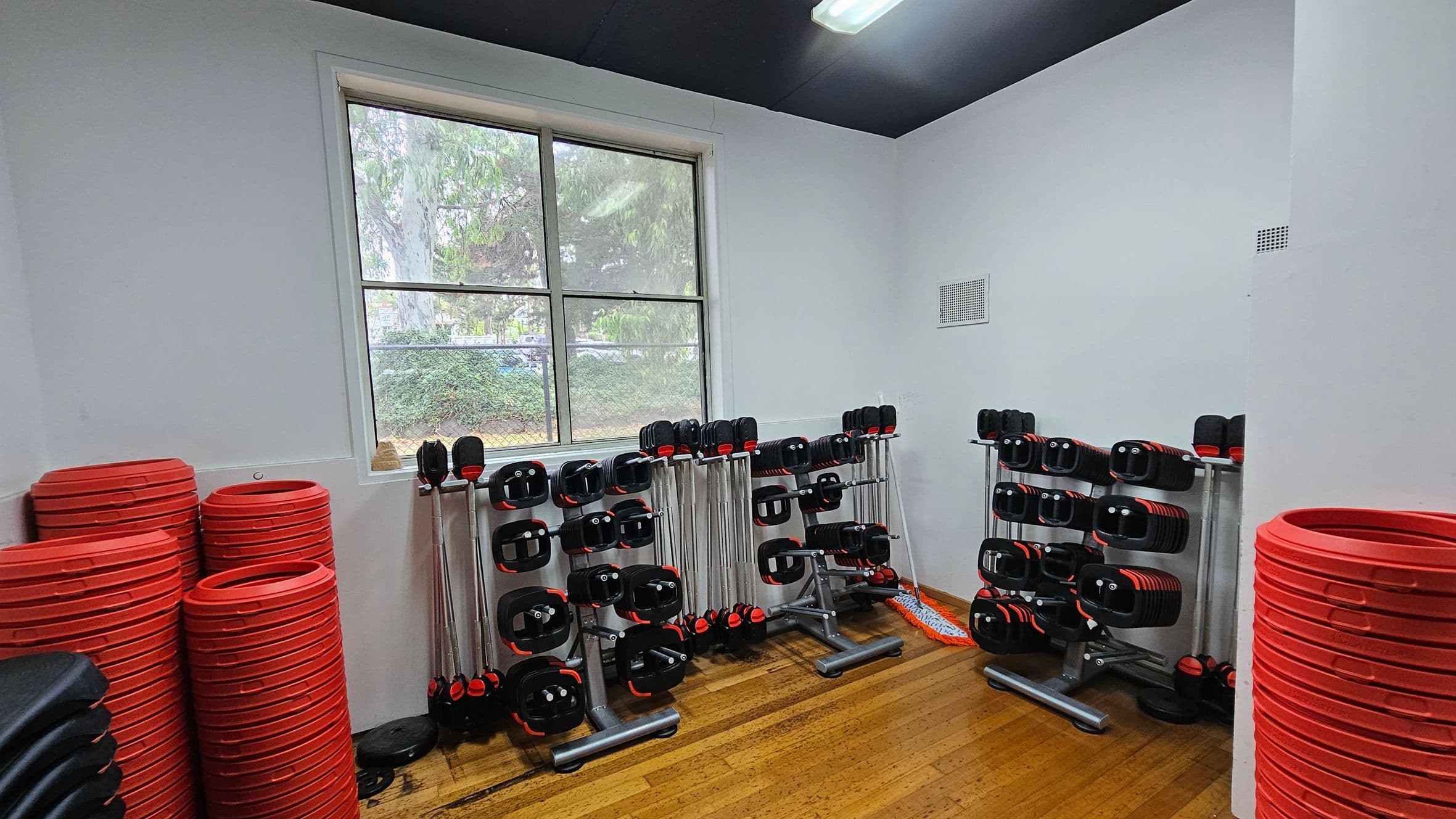 A gym room with wooden flooring and a black ceiling. On the left, there are stacks of red weight plates and black step platforms. In the centre, several racks hold barbells with black and red weights. A window in the background shows greenery outside. On the right, more red weight plates are stacked. A mop is leaning against the wall near the racks.