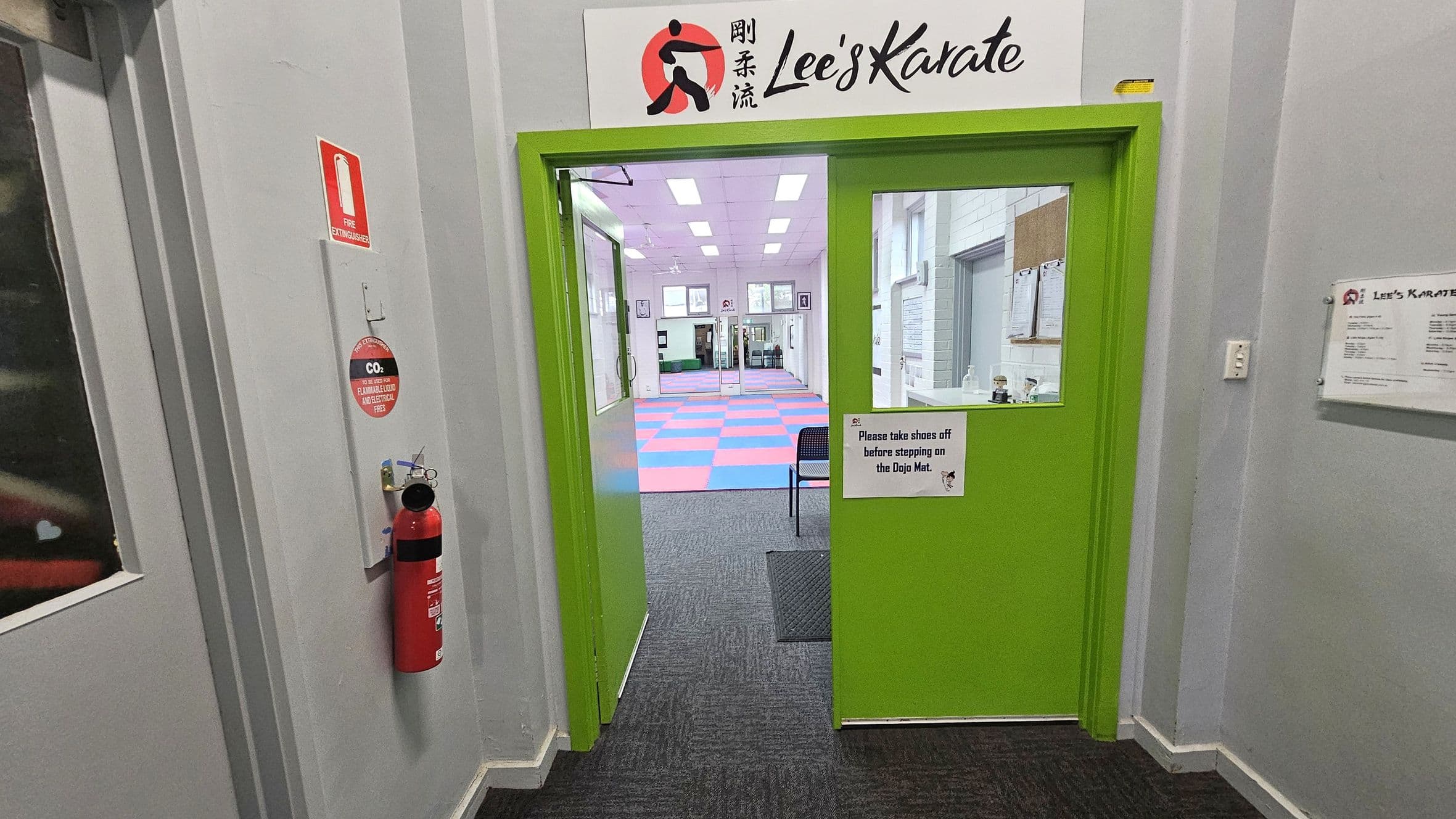 A bright green door with a sign above it reading "Lee's Karate" leads into a room with a colourful checkered mat. A sign on the door requests visitors to remove shoes before stepping on the mat. To the left, a red fire extinguisher is mounted on the wall. The floor is carpeted in dark grey, and the walls are light grey. Inside the room, there are framed pictures and a chair visible. The ceiling has rectangular lights.
