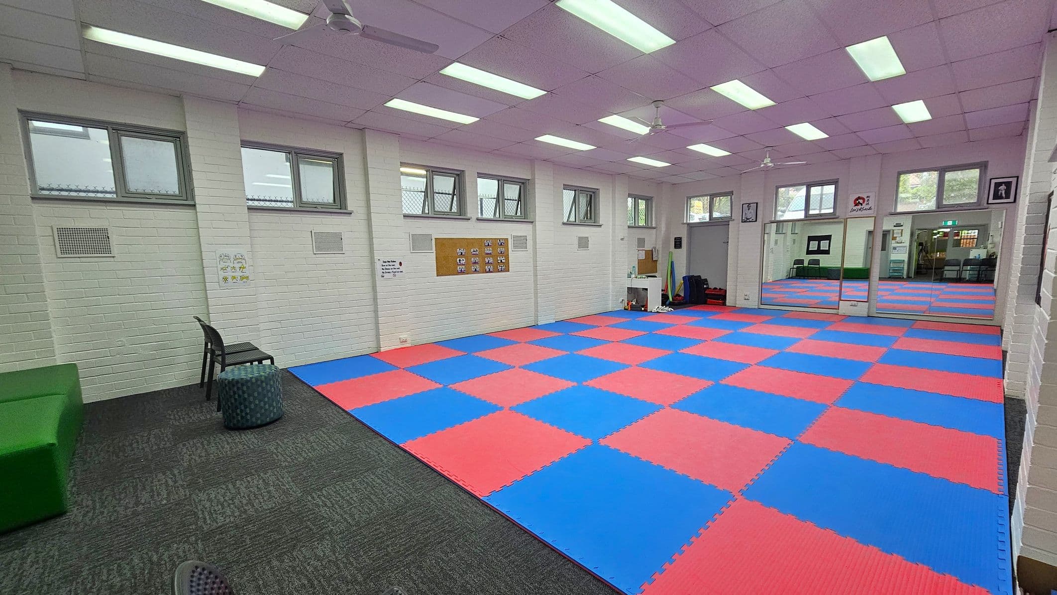 A room with a blue and red checkered mat covering most of the floor. On the left, there is a green bench, a black chair, and a small round stool on a grey carpeted area. The walls are white with several small windows near the ceiling. On the right, there are large mirrors and a doorway leading to another room. The ceiling has fluorescent lights and ceiling fans. Various posters and a bulletin board are on the walls.