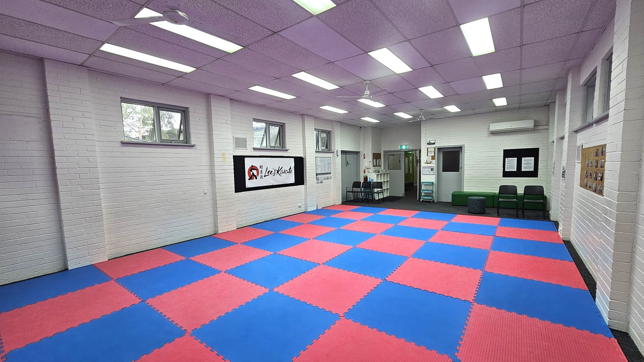 A martial arts studio with a floor covered in interlocking red and blue mats. The walls are white brick with several small windows near the ceiling. On the left wall, there is a sign with text and a logo. The right side has a bulletin board with photos. At the back, there are chairs, a green bench, and a small table. The ceiling is purple with fluorescent lights and a ceiling fan.