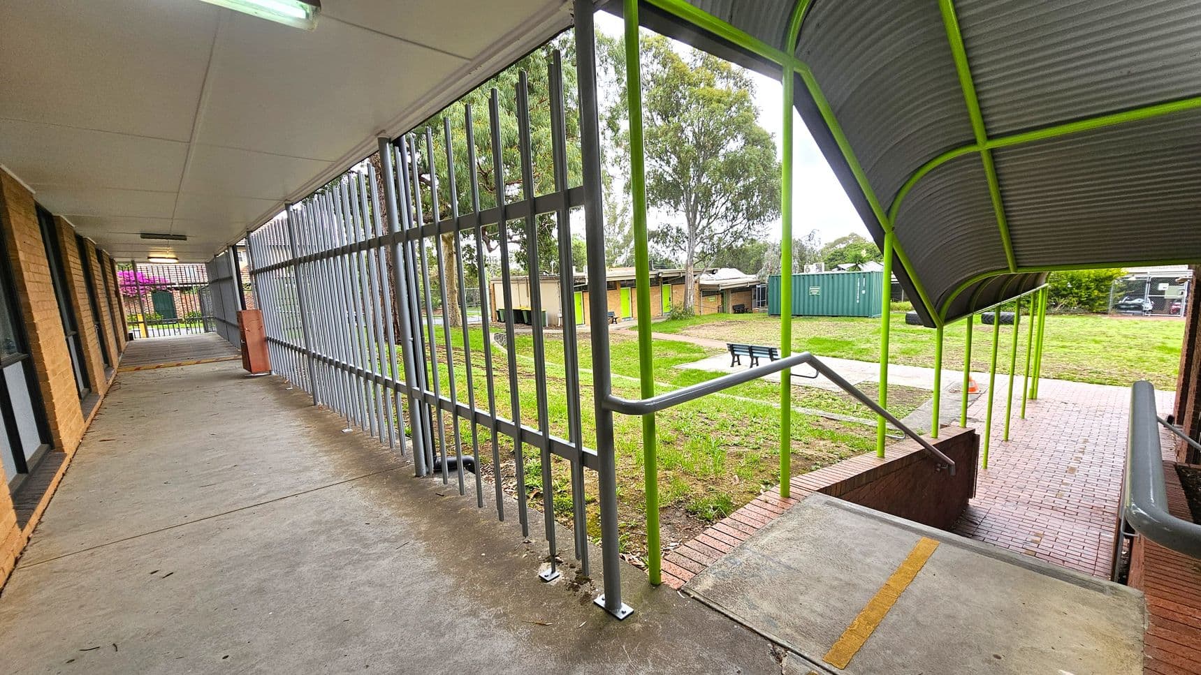 A covered walkway with a concrete floor and a white ceiling on the left, featuring brick walls and windows. Metal bars form a fence along the side. On the right, a ramp with a handrail leads down to a brick path. The roof above the ramp is supported by green poles. In the background, there is a grassy area with trees, a green shipping container, and a few small buildings. A bench is visible on the grass.