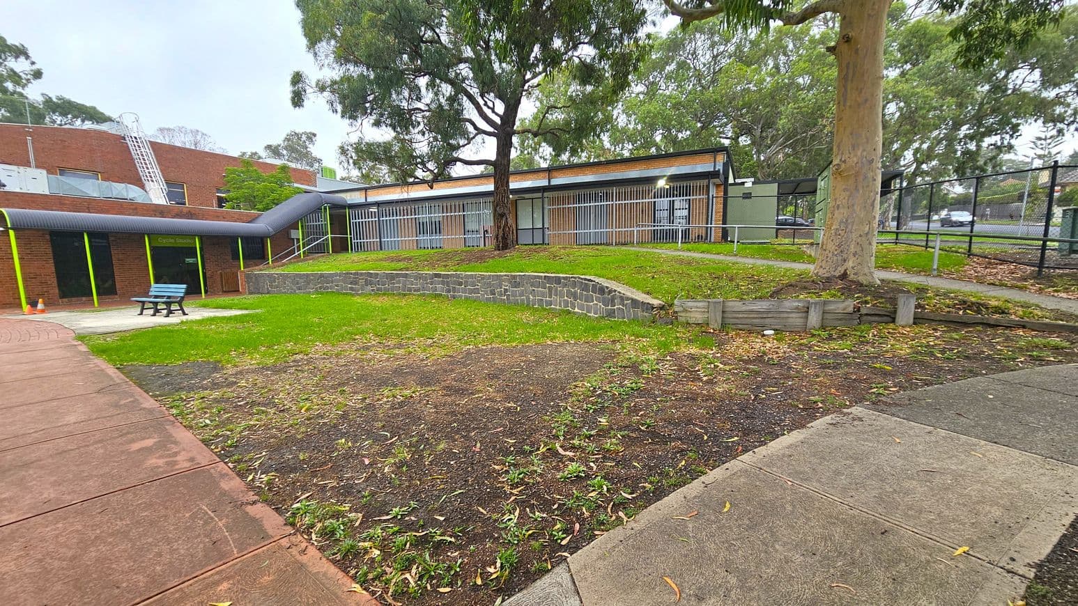 A paved walkway leads to a brick building with large windows and metal bars. On the left, a blue bench sits on a concrete area near a "Cycle Studio" sign. A large tree stands in the centre, surrounded by grass and a low stone wall. The right side features a fenced area with more trees and a path. The ground is a mix of grass and bare patches, with scattered leaves.
