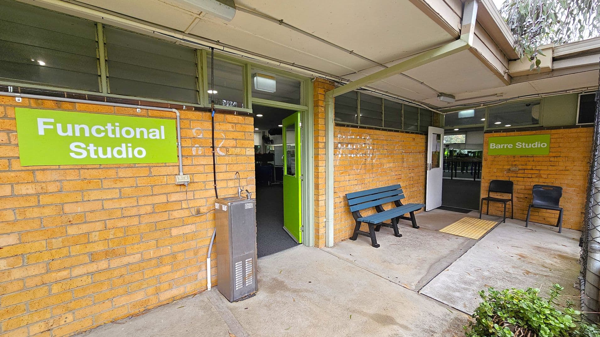 A brick building with two entrances. On the left, a green sign reads "Functional Studio" above a green door, with a water fountain nearby. On the right, a sign says "Barre Studio" above a white door, with two chairs beside it. A blue bench is positioned between the entrances. The floor is concrete, and the ceiling is light-coloured with visible piping. A small bush is in the foreground near the right entrance.