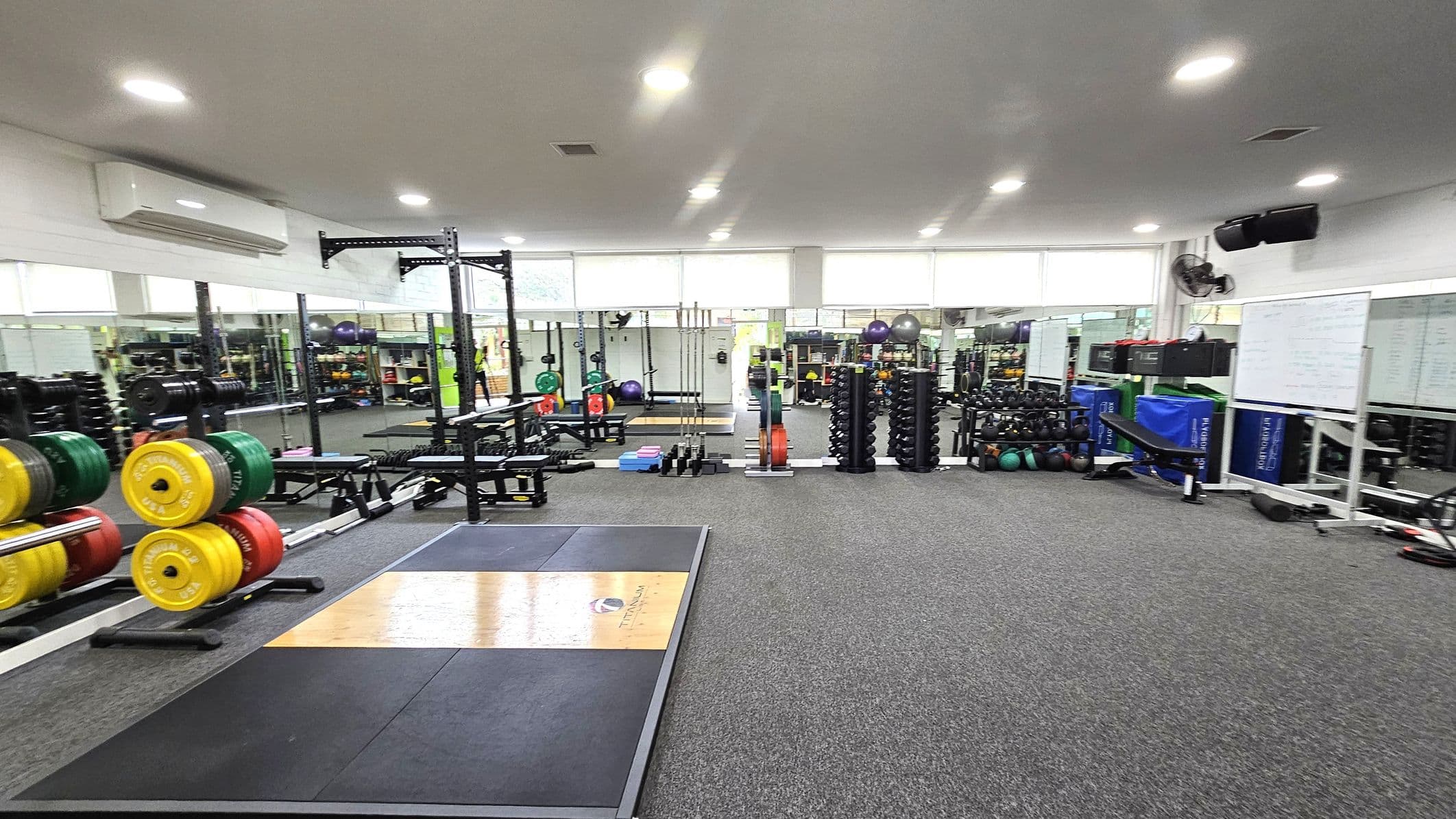 A spacious gym with a grey carpeted floor and a white ceiling with recessed lighting. On the left, there are weightlifting platforms with colourful weight plates stacked on racks. In the centre, there are various exercise equipment, including benches and dumbbells. The right side features more weights, exercise balls, and a whiteboard. Large mirrors line the walls, reflecting the equipment and adding depth to the space.