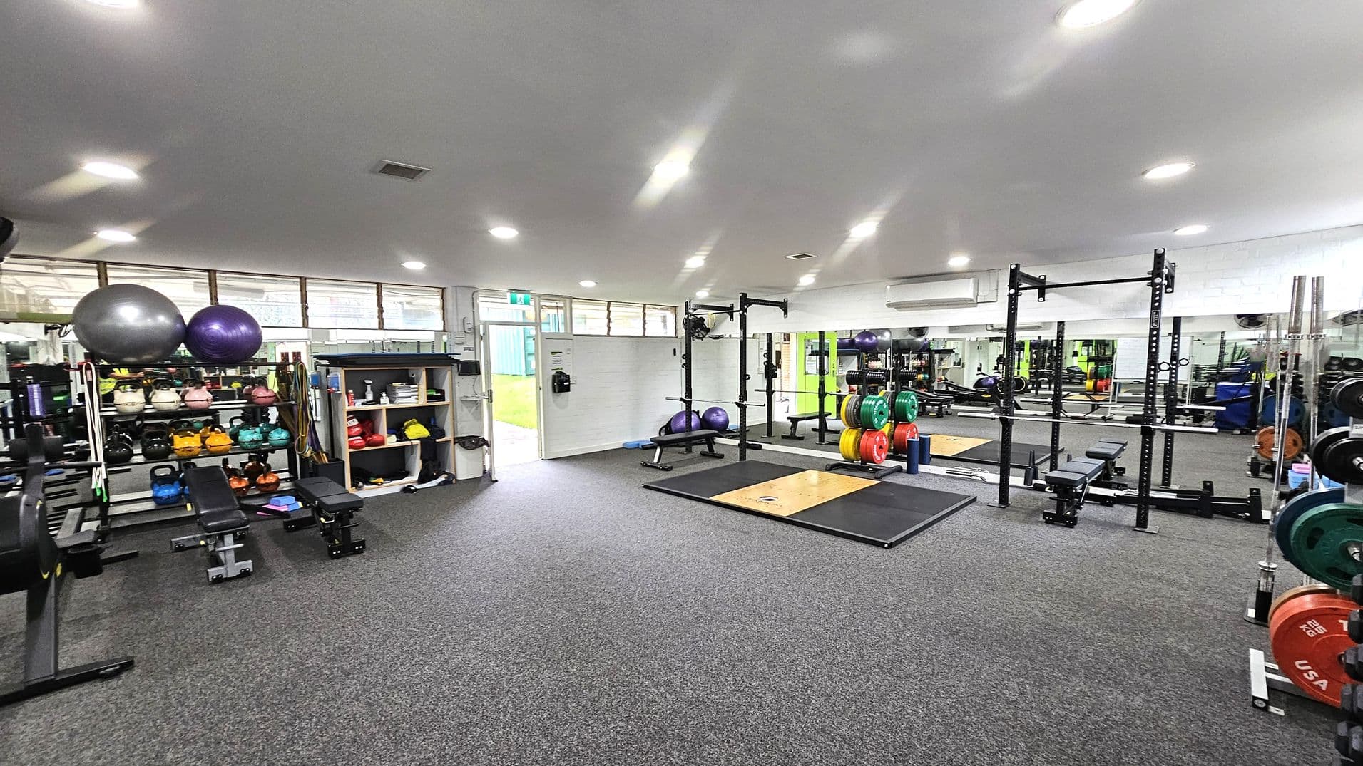 A gym with a grey carpeted floor and a white ceiling with recessed lights. On the left, there are shelves with kettlebells, medicine balls, and exercise balls. In the centre, a squat rack with colourful weight plates and a lifting platform is visible. The right side features more weightlifting equipment and mirrors along the wall. An open door at the back leads outside, allowing natural light to enter.