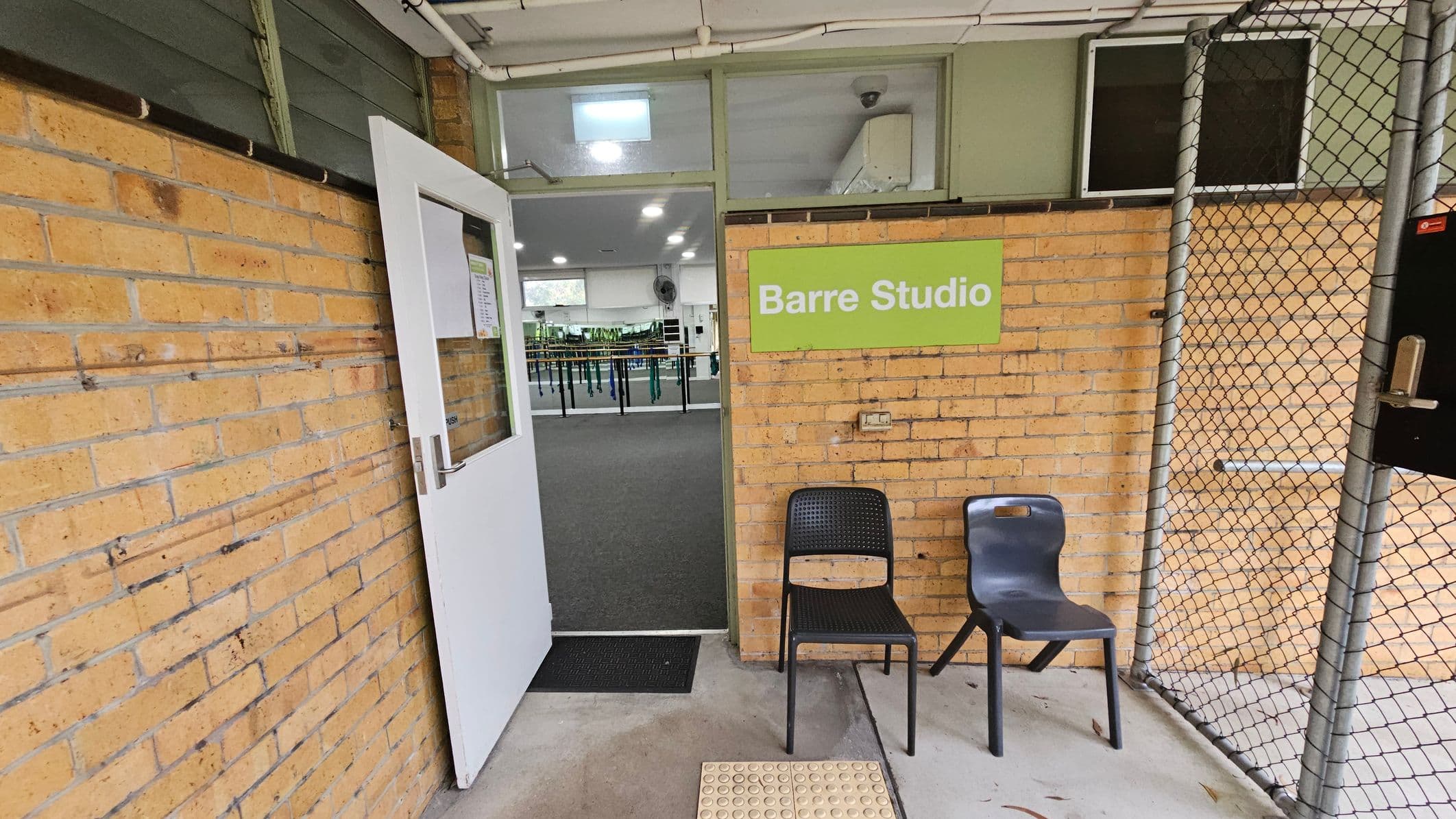 A brick wall with a white door slightly open, leading into a room with a grey carpet and exercise equipment. A green sign on the wall reads "Barre Studio". Two chairs, one black and one grey, are placed outside the door. A chain-link fence is on the right side. The ceiling is white with visible pipes, and the floor is concrete with a textured mat near the entrance.