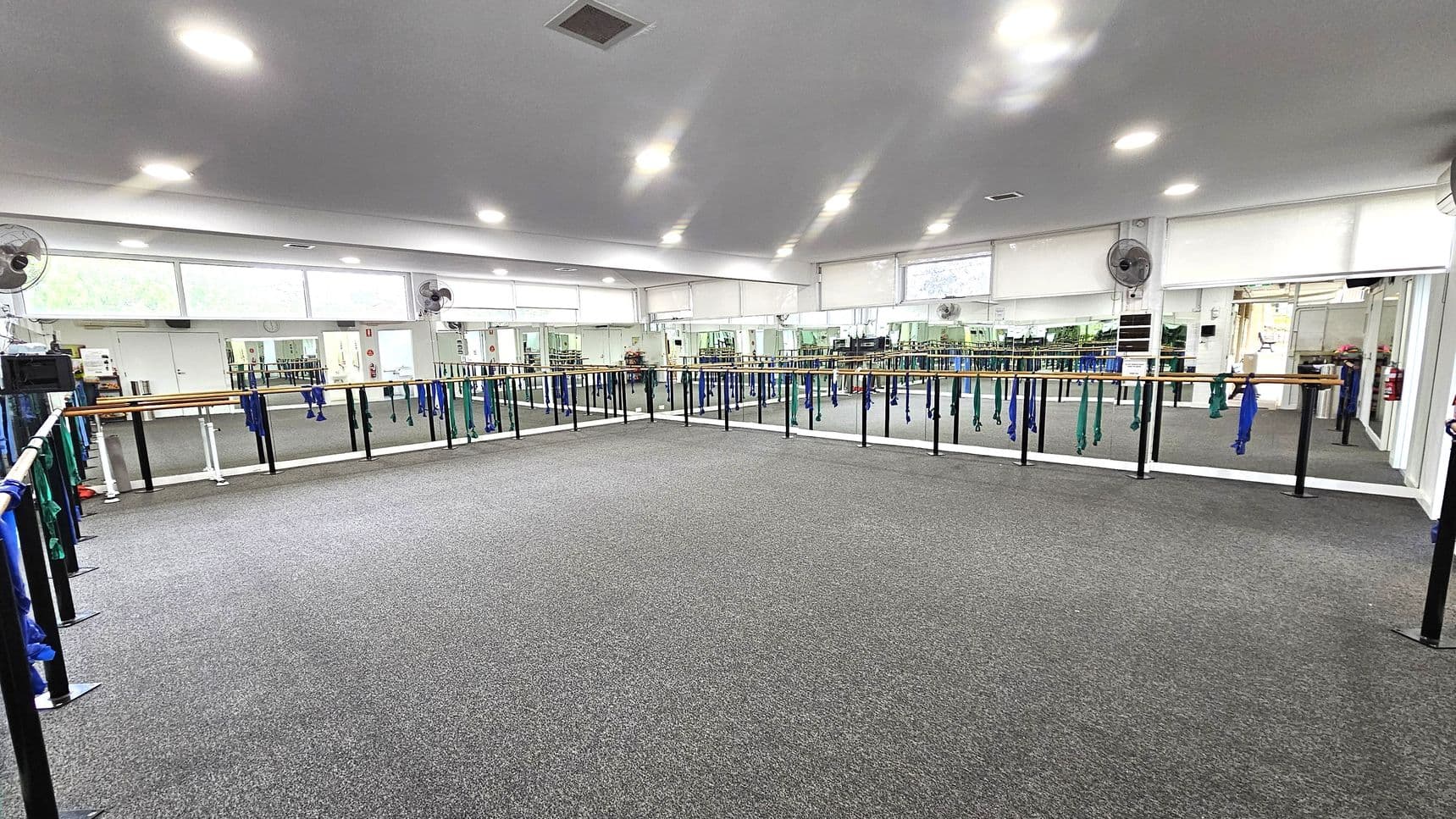 A spacious dance studio with grey carpeted flooring. The room features mirrored walls on the left and right, with ballet barres running along them. Blue and green resistance bands hang from the barres. The ceiling is white with recessed lighting, and there are several wall-mounted fans. Large windows above the mirrors allow natural light to enter. The room is empty, providing ample space for movement.
