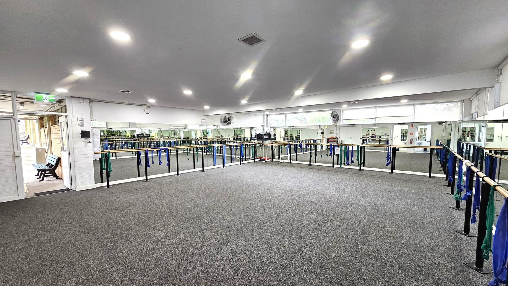 A spacious dance studio with grey carpeted flooring and a white ceiling with recessed lights. The walls are lined with mirrors, and ballet barres run along them, adorned with blue and green resistance bands. On the left, there's an open door leading to a hallway with a bench. The room is well-lit, and there are ceiling fans above the mirrors.