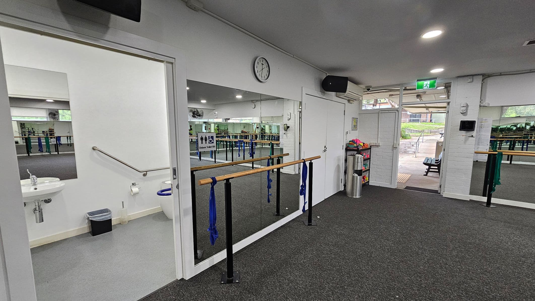 A dance studio with a grey carpeted floor. On the left, there's an open door leading to a bathroom with a sink, toilet, and handrail. In the centre, a mirrored wall with a wooden ballet barre attached, adorned with blue fabric. A clock is mounted above the mirror. On the right, an open door leads outside, showing a paved area with benches. A shelf with colourful items and a silver bin is near the door. The ceiling has recessed lighting.