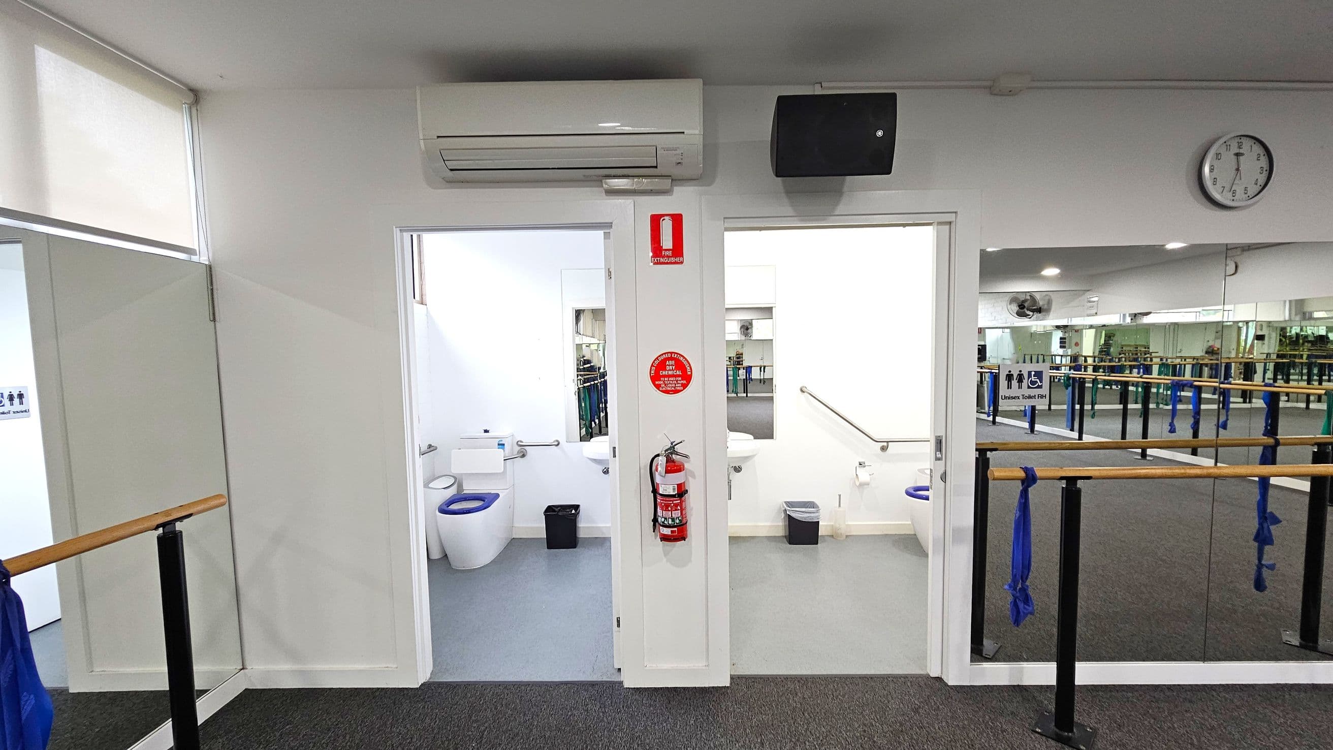 A room with two open doors leading to toilets, each with a white toilet and a small bin. Above the doors, an air conditioning unit and a speaker are mounted. A fire extinguisher is attached to the wall between the doors. On the right, a wall clock shows the time. The foreground features a mirrored wall with ballet barres and blue fabric tied to them. The floor is carpeted, and the ceiling has recessed lighting.