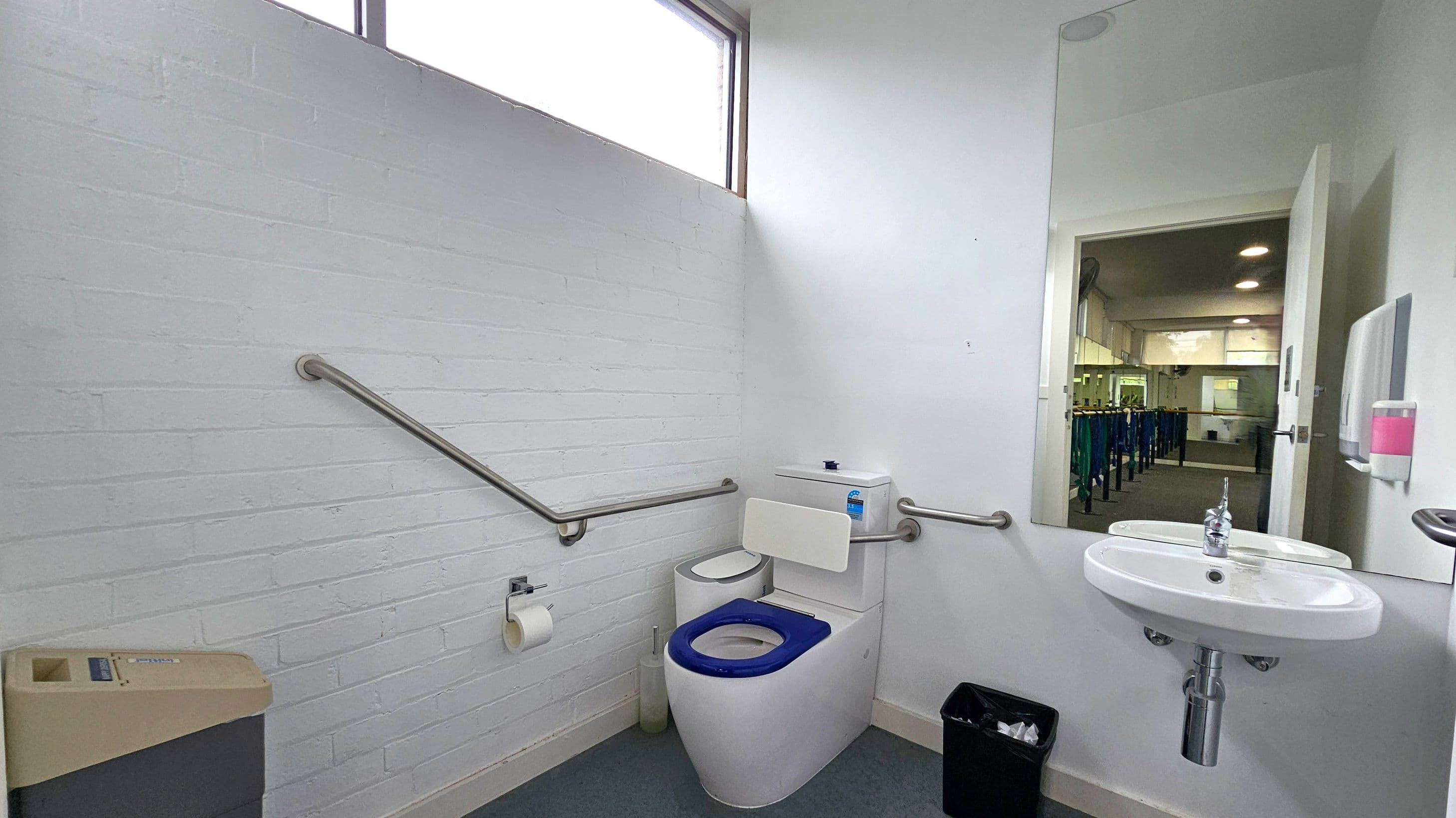 A bathroom with white walls and a grey floor. On the left, there is a beige bin and a metal handrail along the wall. In the centre, a toilet with a blue seat and a white backrest is positioned against the wall, with a toilet roll holder nearby. On the right, a white sink with a chrome tap is mounted below a large mirror reflecting a room with tables and chairs. A soap dispenser is mounted on the wall next to the sink. A window is visible at the top of the left wall.