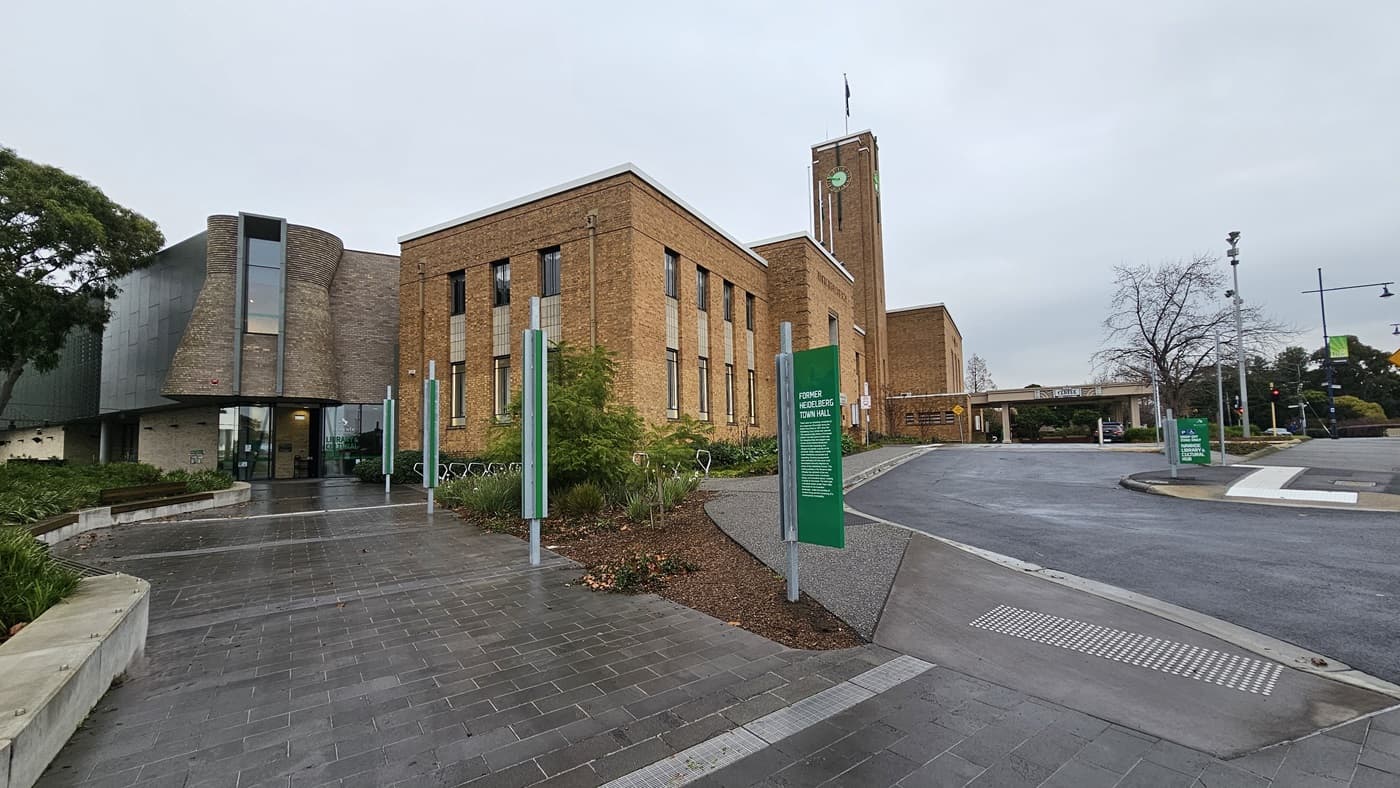 On the left, there is a modern building with a curved glass facade and a garden with green shrubs and trees. In the middle, there are several green signs with white text and a pathway made of grey tiles. On the right, a large brick building with tall windows and a clock tower. The foreground features a grey tiled floor and a wet asphalt road with a zebra crossing.