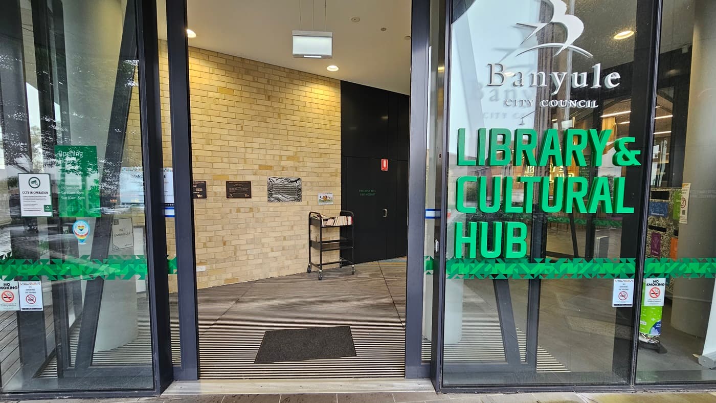 On the left, there is a glass door with a green stripe and various stickers. In the middle, a brick wall with a small table holding brochures and a black mat on the floor. On the right, another glass door with the text "Banyule City Council" and "Library & Cultural Hub" in green. The floor is a combination of wooden planks and a ribbed mat at the entrance. The ceiling is white with recessed lighting and a hanging light fixture.